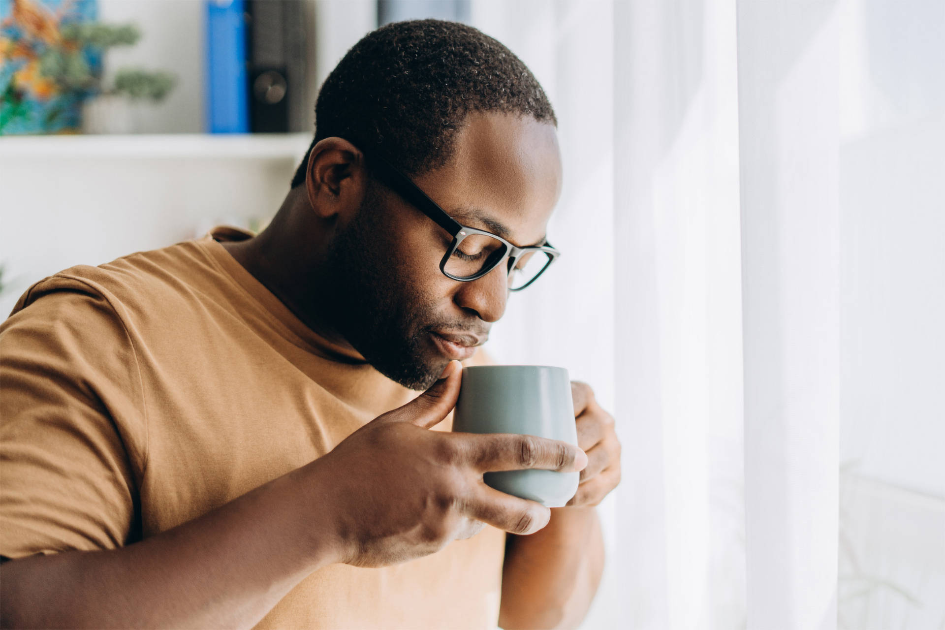 Mann mit Brille und braunem T-Shirt trinkt aus einer Tasse, während er am Fenster steht.
