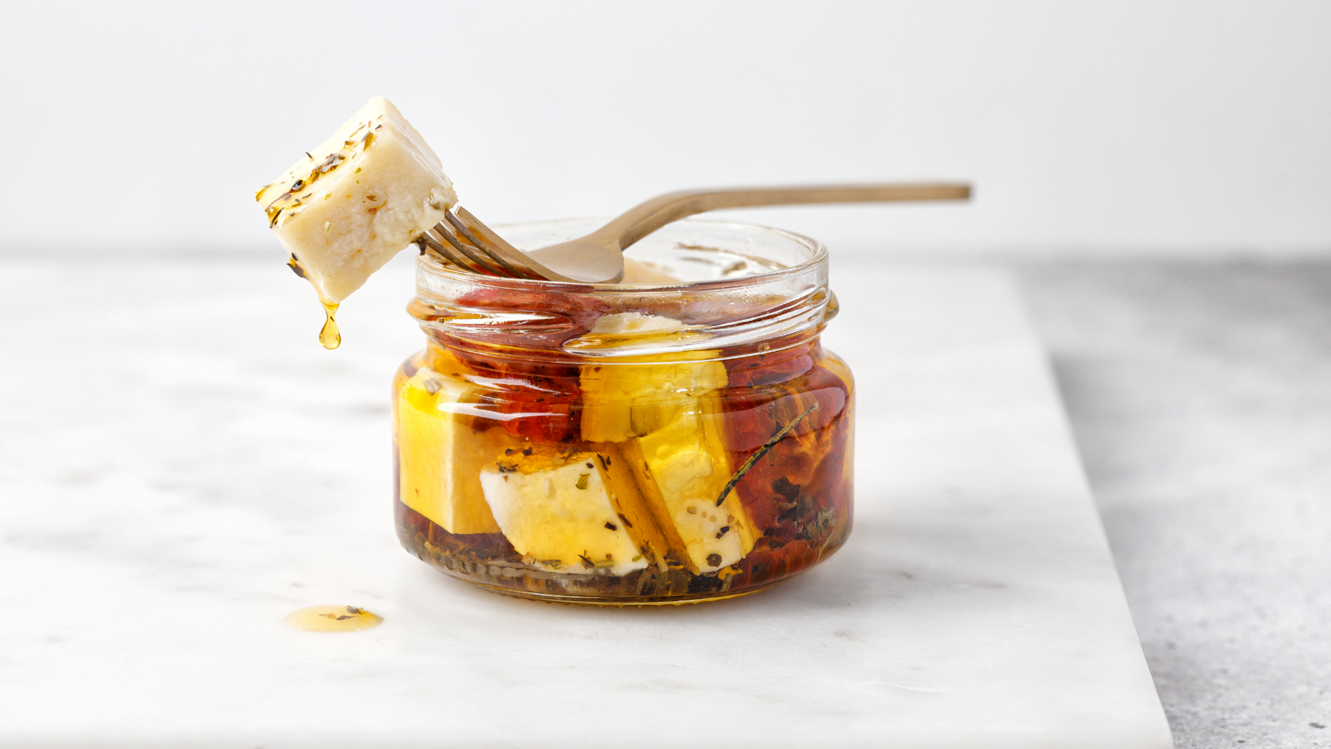 Eingelegter Feta/Hirtenkäse im Glas mit Gabel auf Marmor.