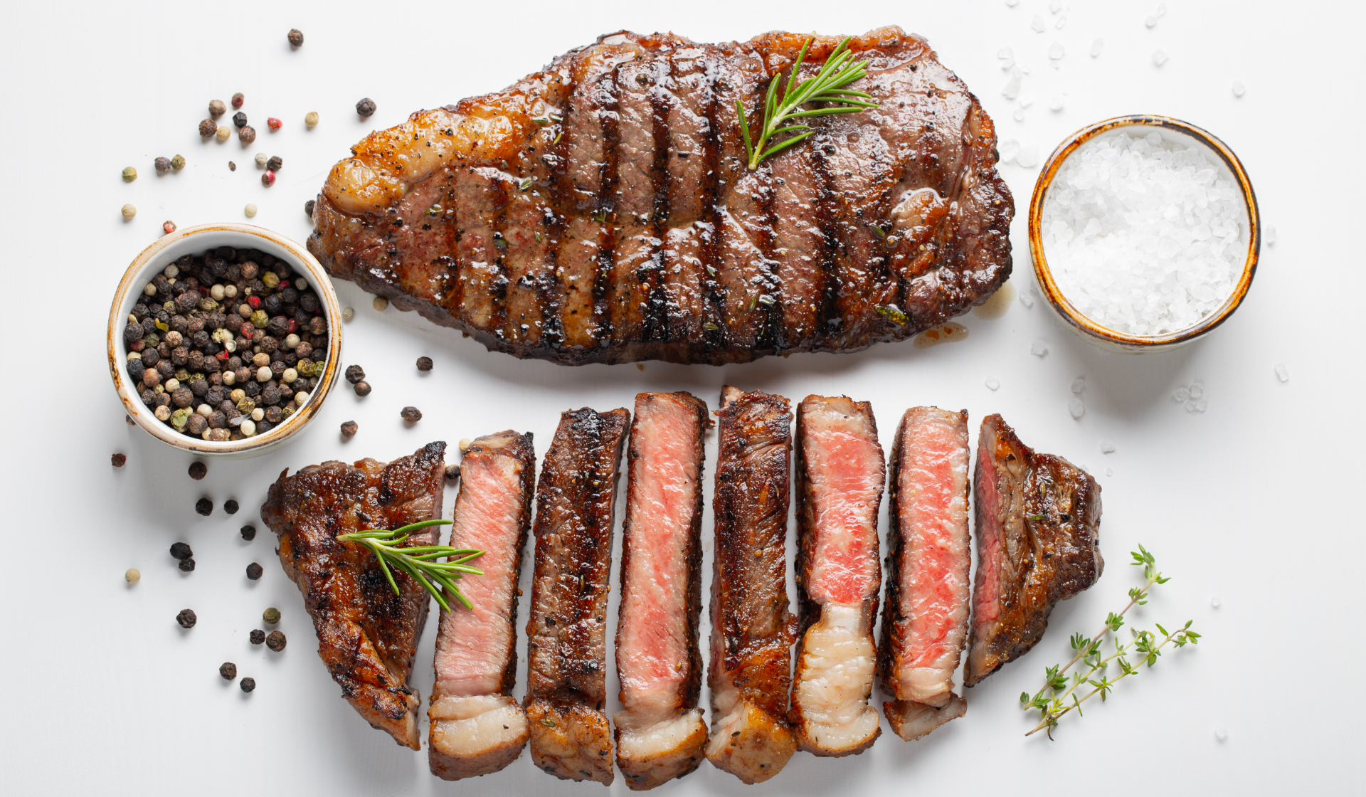Zwei gebratene Rindfleischstücke und daneben eine Schüssel mit ganzen Pfefferkörnern und eine Schüssel mit groben Salz.