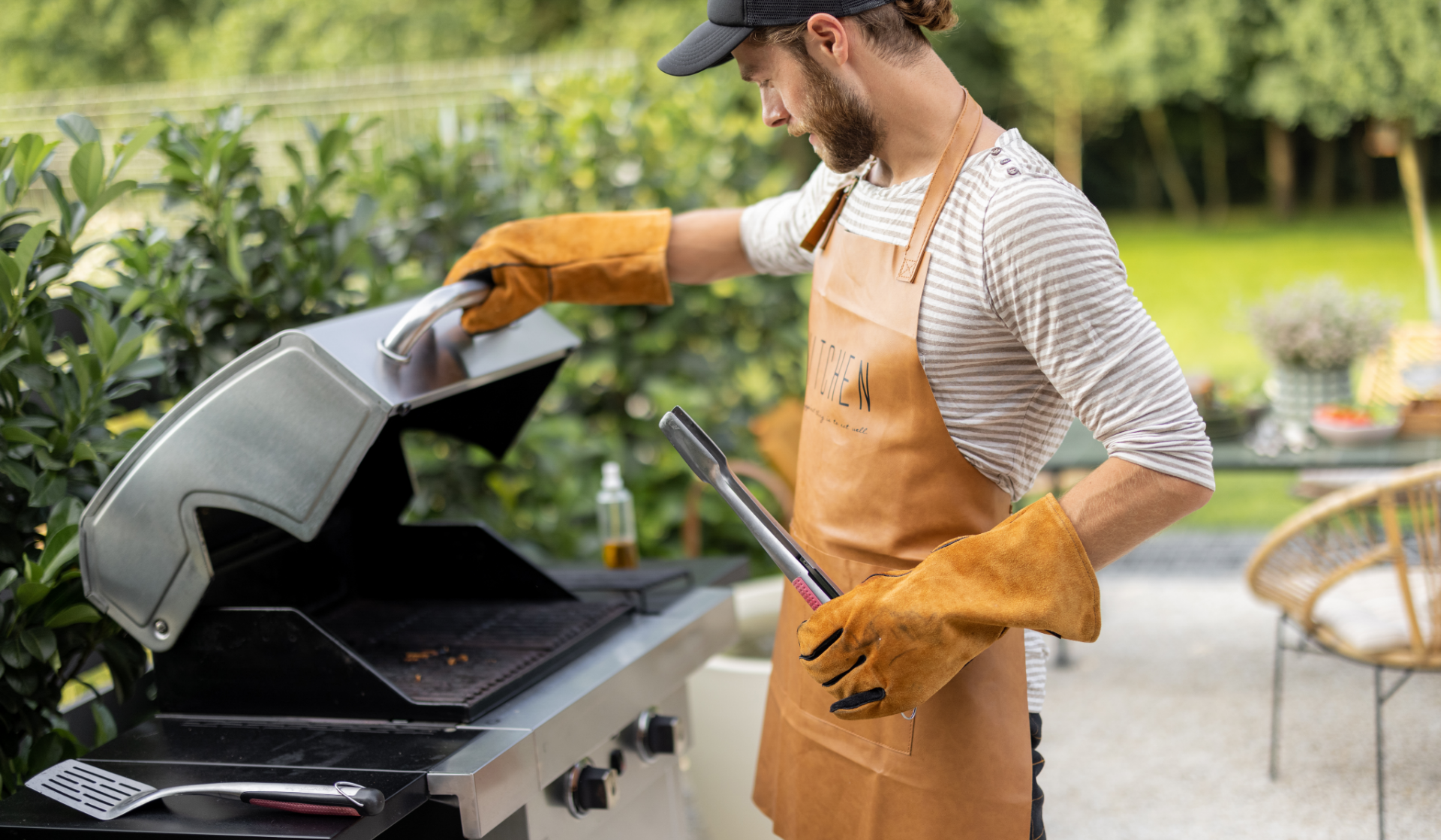 Mann mit Grillhandschuhen, Schürze, Baseballcap und Grillzange in der Hand steht auf der Terrasse am Grill.