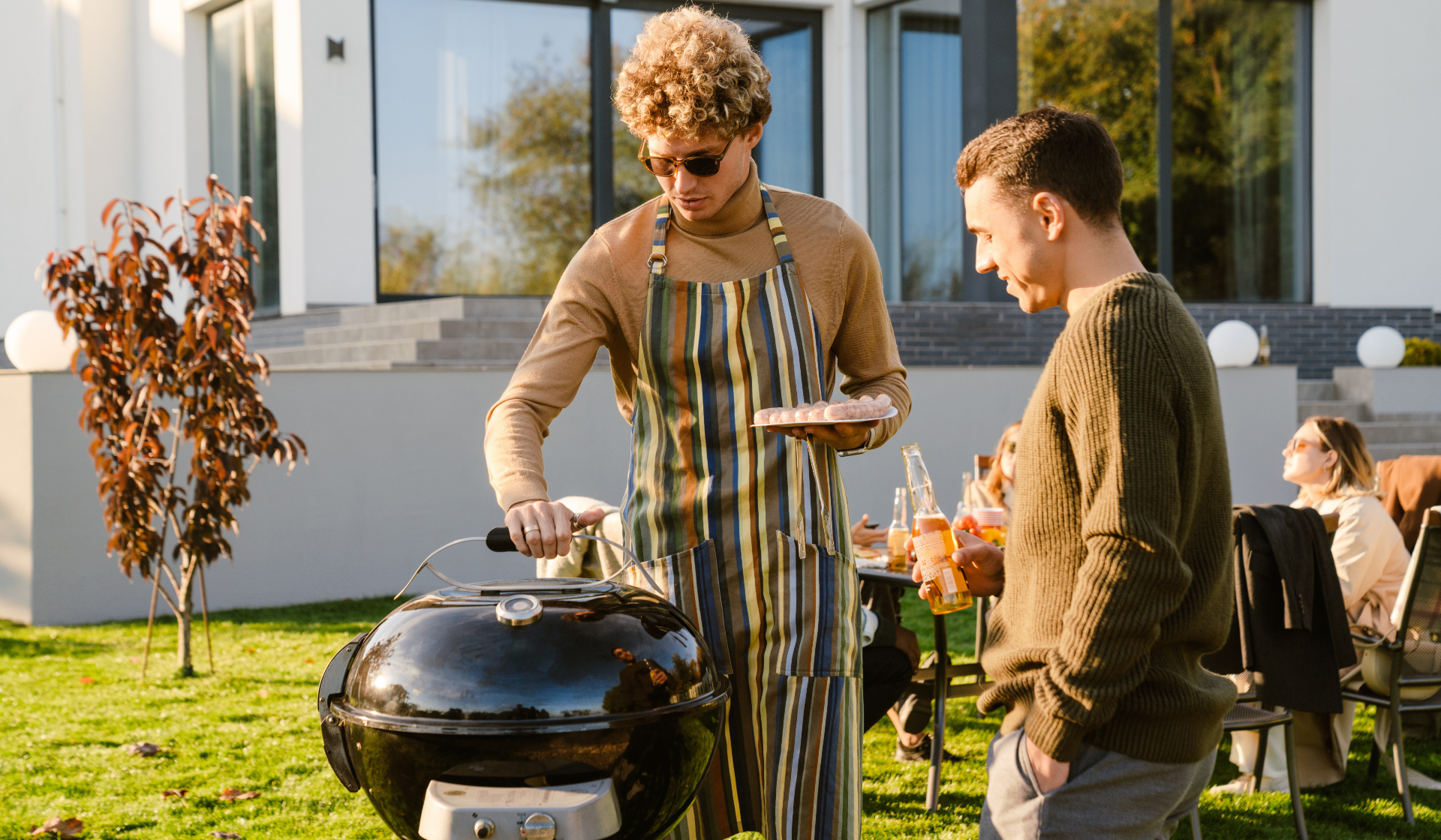 Zwei Männer stehen draußen am Grill, einer mit Sonnenbrille, Schürze und einem Teller mit ungegrillten Würstchen.