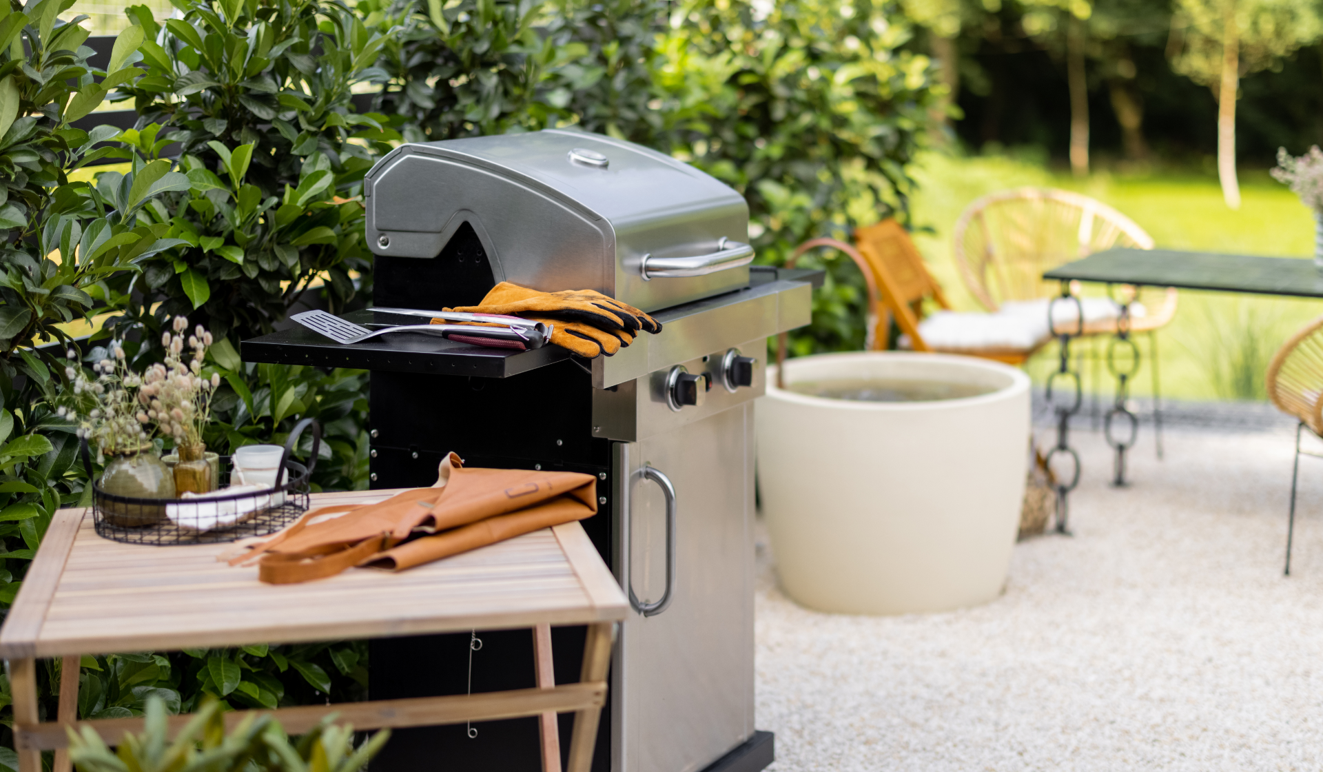 Kleiner Holztisch mit gefalteter Schürze darauf, daneben Gasgrill mit Grillhandschuhen und Grillbesteck, dahinter Hecken