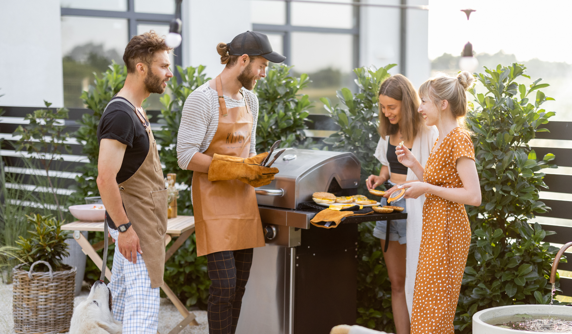 Zwei Männer in Grillschürze und zwei Frauen stehen um einen Gasgrill und bereiten Hamburger vor.
