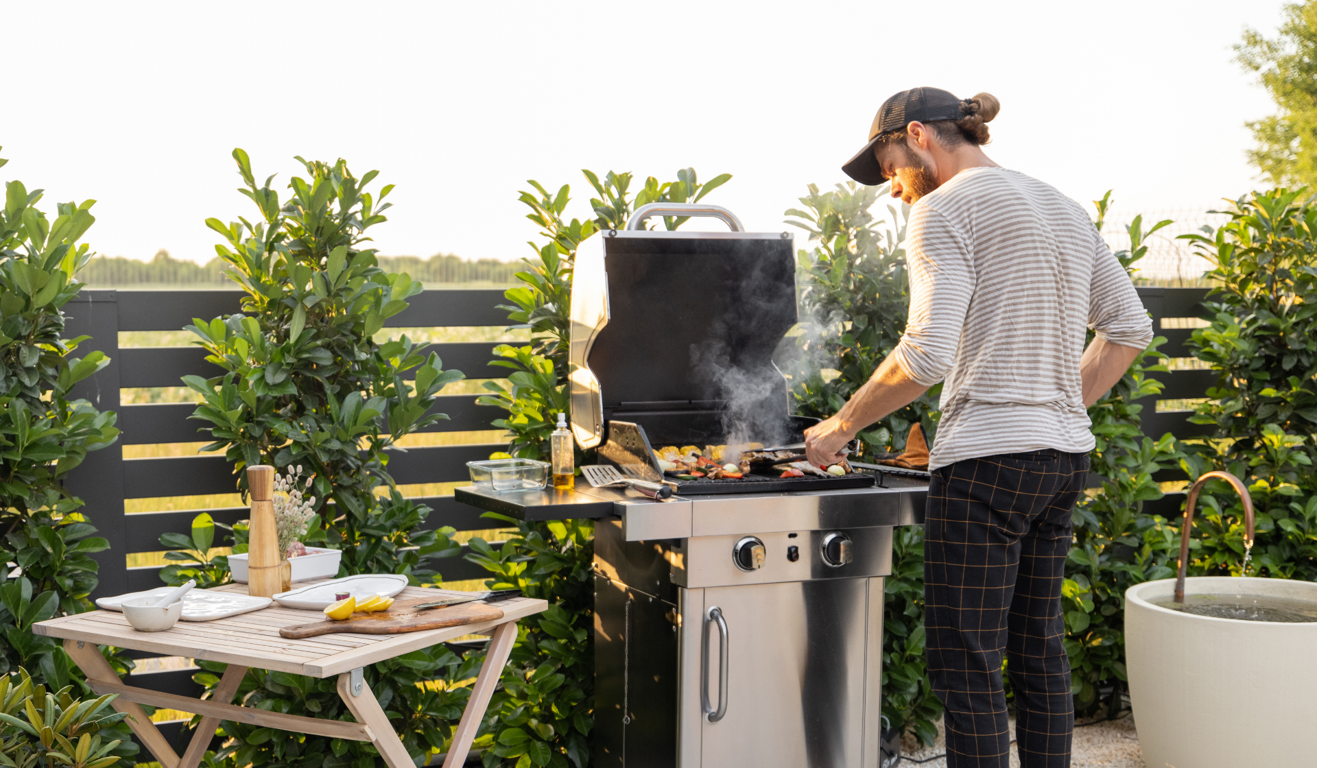 Mann in gestreiftem Shirt, karierter Hose und Schildmütze steht mit Grillzange am mit Gemüse belegten Gasgrill.