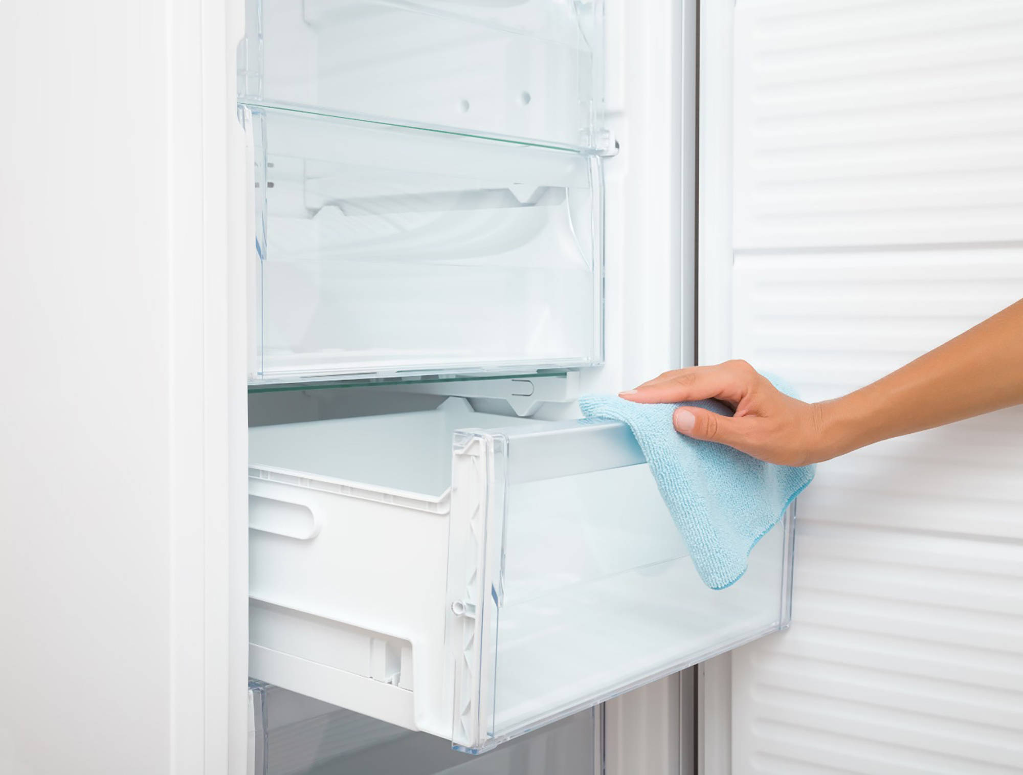 Jemand wischt mit einem blauen Mikrofasertuch die transparente Gefrierschublade eines weißen, sauberen Kühlschranks aus.