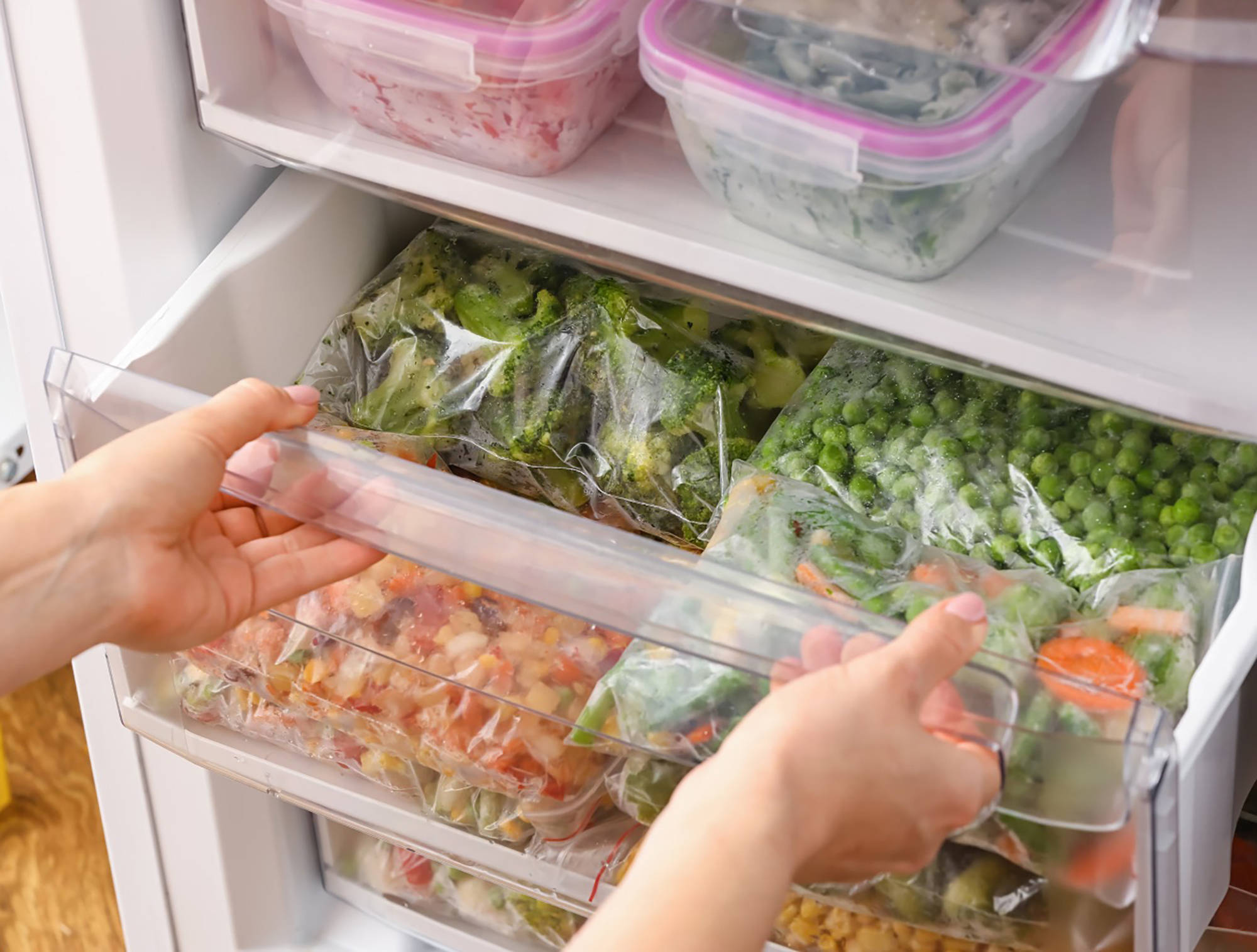Person öffnet eine Gefrierschublade mit gefrorenem Gemüse in Plastikbeuteln, darunter Brokkoli, Erbsen und Mischgemüse.