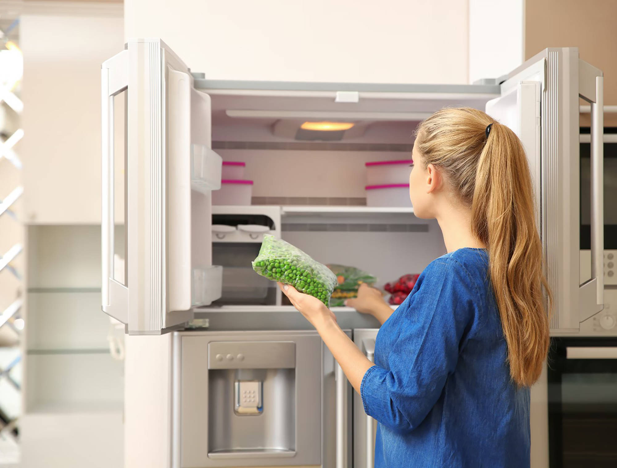Frau legt eine Tüte gefrorene Erbsen in einen doppeltürigen Gefrierschrank in einer modernen Küche.