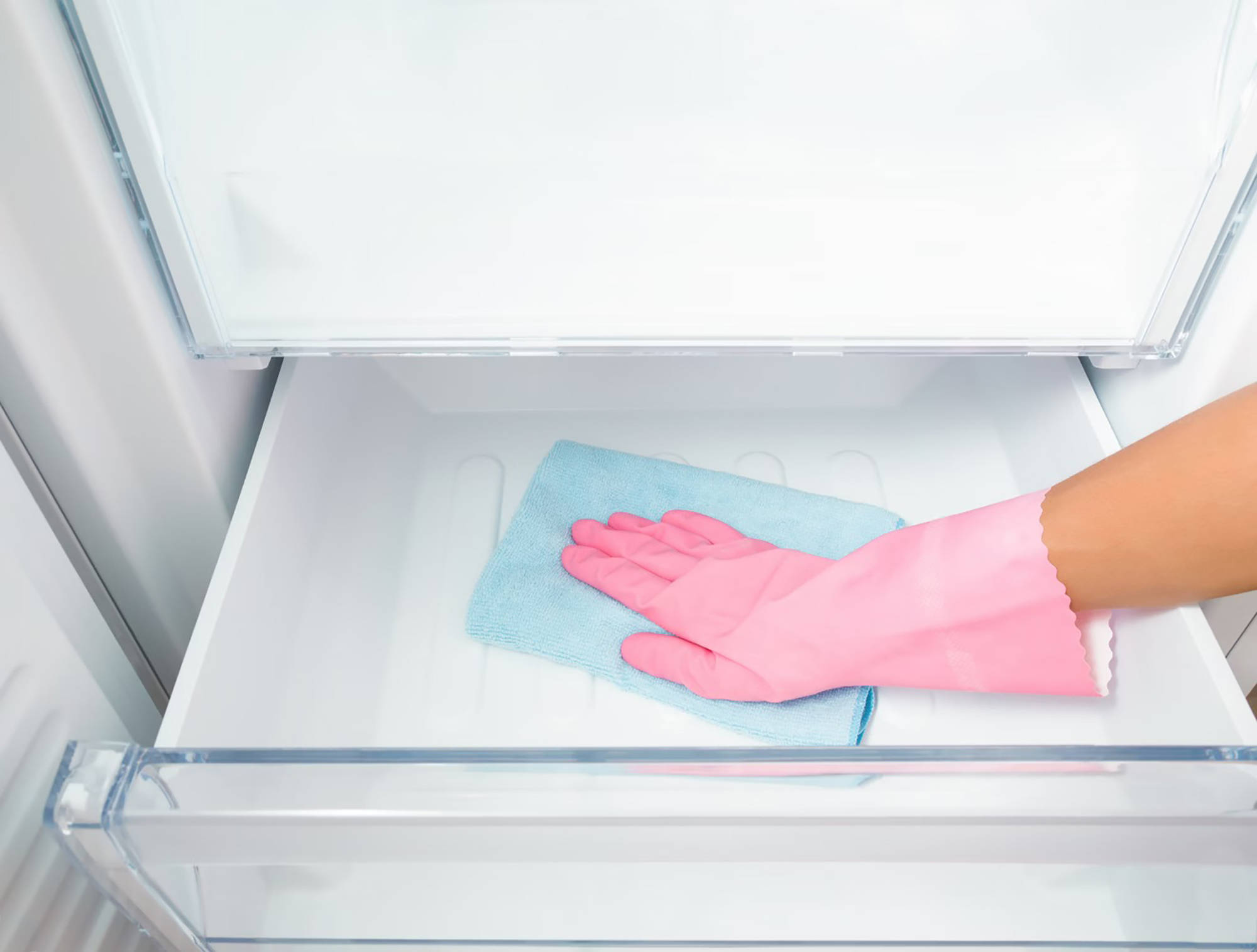 Person mit rosa Gummihandschuhen reinigt ein leeres Gefrierfrach mi einem blauen Mikrofasertuch.