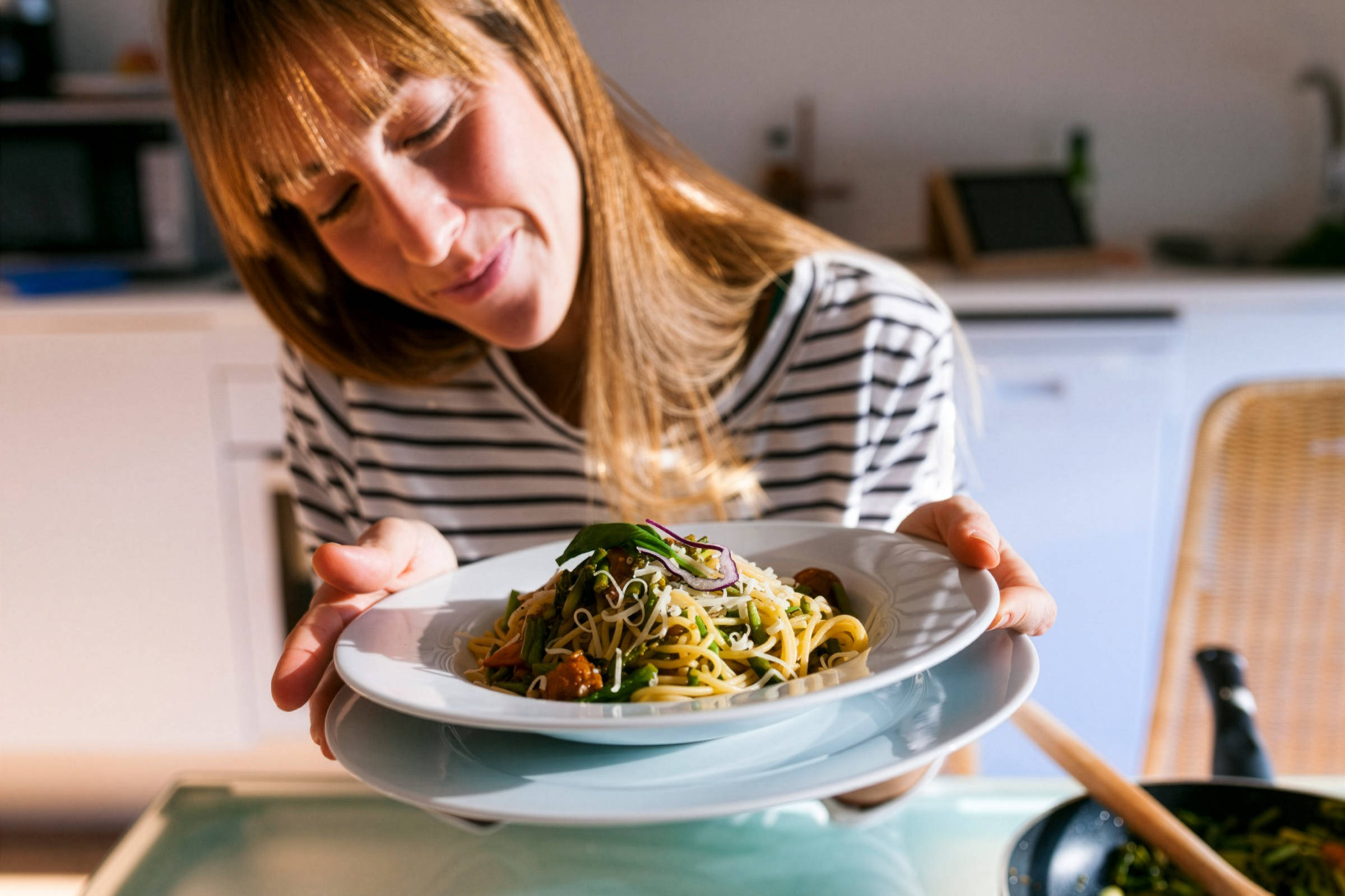 Frau präsentiert stolz einen Teller Spaghetti mit Gemüse und Käse in einer hellen Küche.