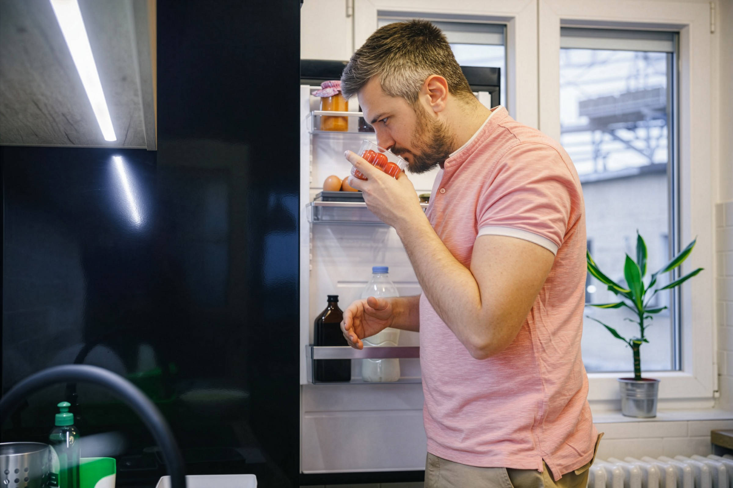 Mann in rosa Polohemd riecht an Tomaten vor offenem Kühlschrank.