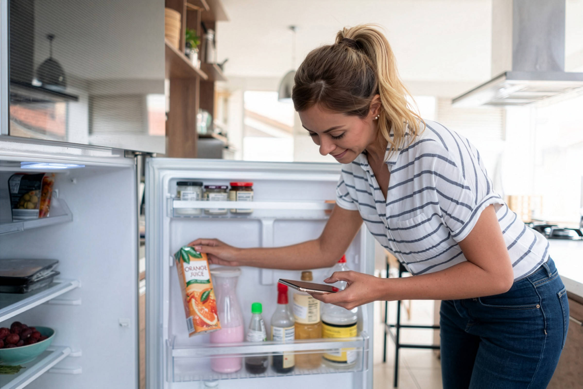  Frau sortiert Lebensmittel im Kühlschrank und schreibt eine Einkaufsliste auf Smartphone