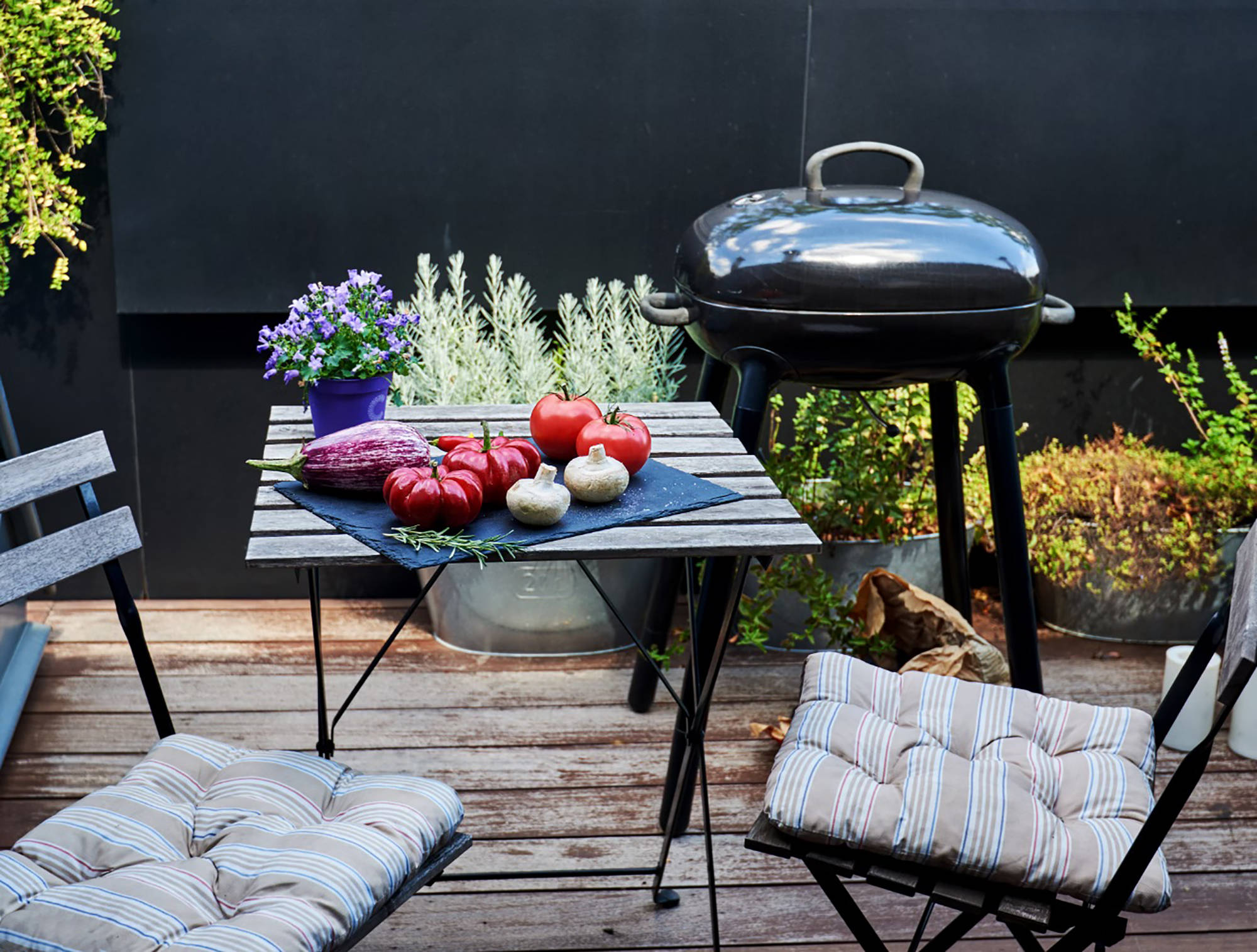 Gemüseauswahl mit Tomaten, Aubergine und Pilzen auf Holztisch vor Kugelgrill auf bepflanzter Terrasse.