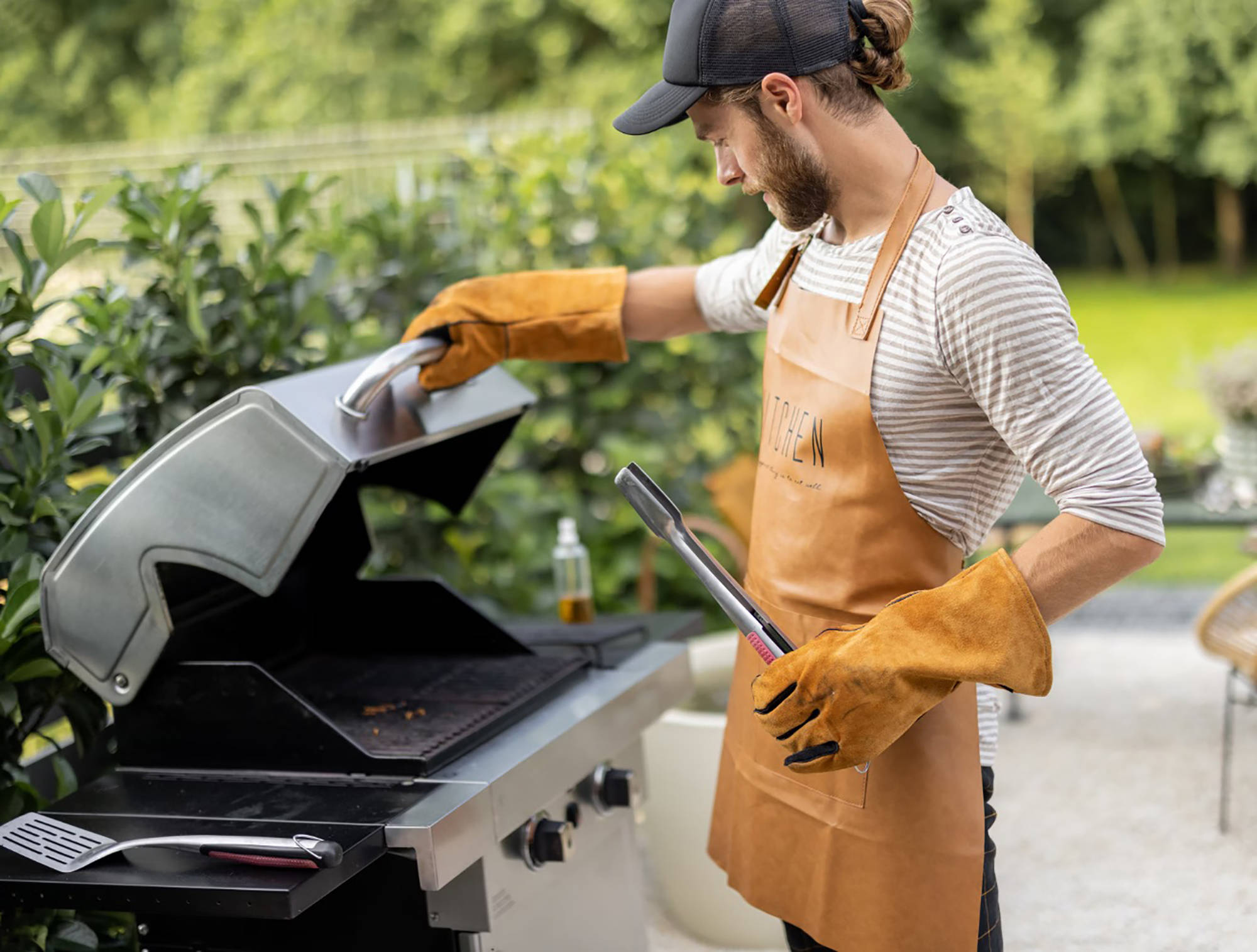 Mann mit Lederhandschuhen und Schürze öffnet Gasgrilldeckel im Garten, Grillzange in der Hand, bereit zum Grillen.