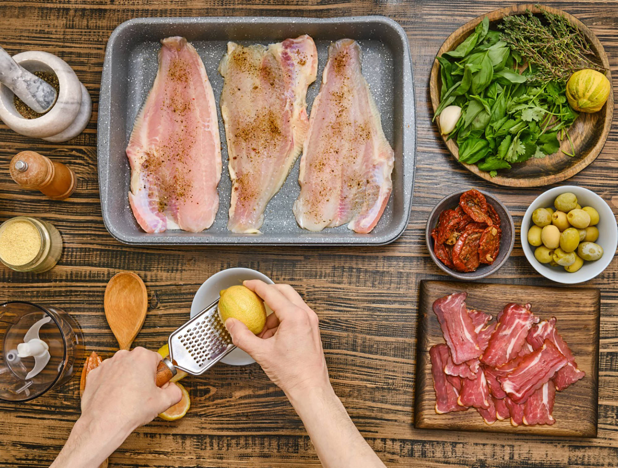 Roh marinierter Fisch in Auflaufform, umgeben von Kräutern, Oliven, getrockneten Tomaten und geriebener Zitrone auf Holz