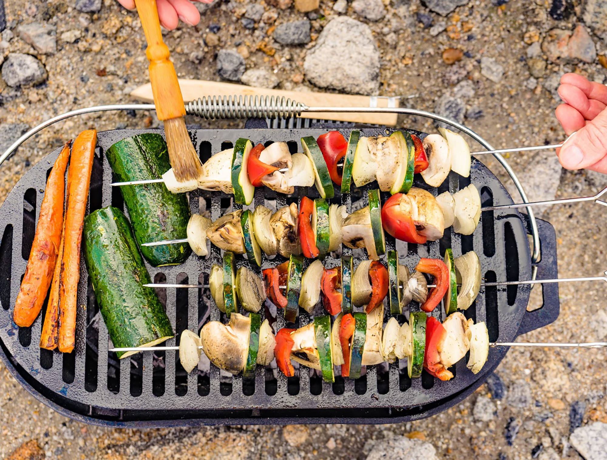 Gemüsespieße mit Paprika, Zucchini, Zwiebeln und Pilzen auf Grillrost, Möhren daneben, Marinade wird aufgetragen.