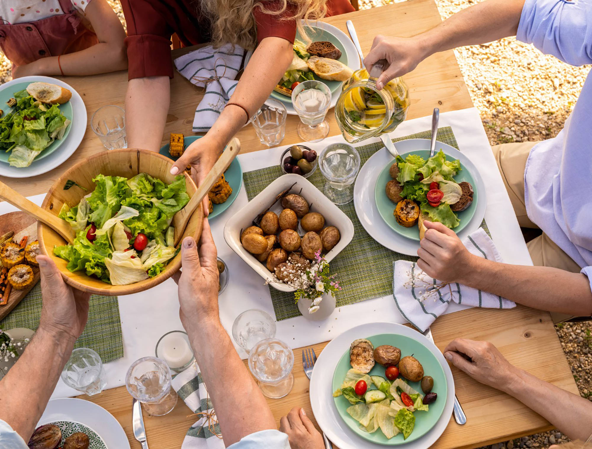 Sommerliche Tischszene mit Grillgemüse, Salat, Kartoffeln und Brot, mehrere Hände greifen zu, Wasser mit Zitrone wird ei