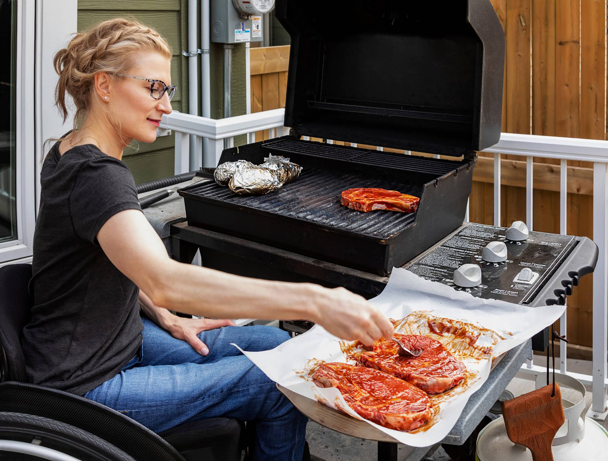 Frau im Rollstuhl grillt mariniertes Fleisch auf Gasgrill, bereitet weiteres Fleisch auf Pergamentpapier vor.