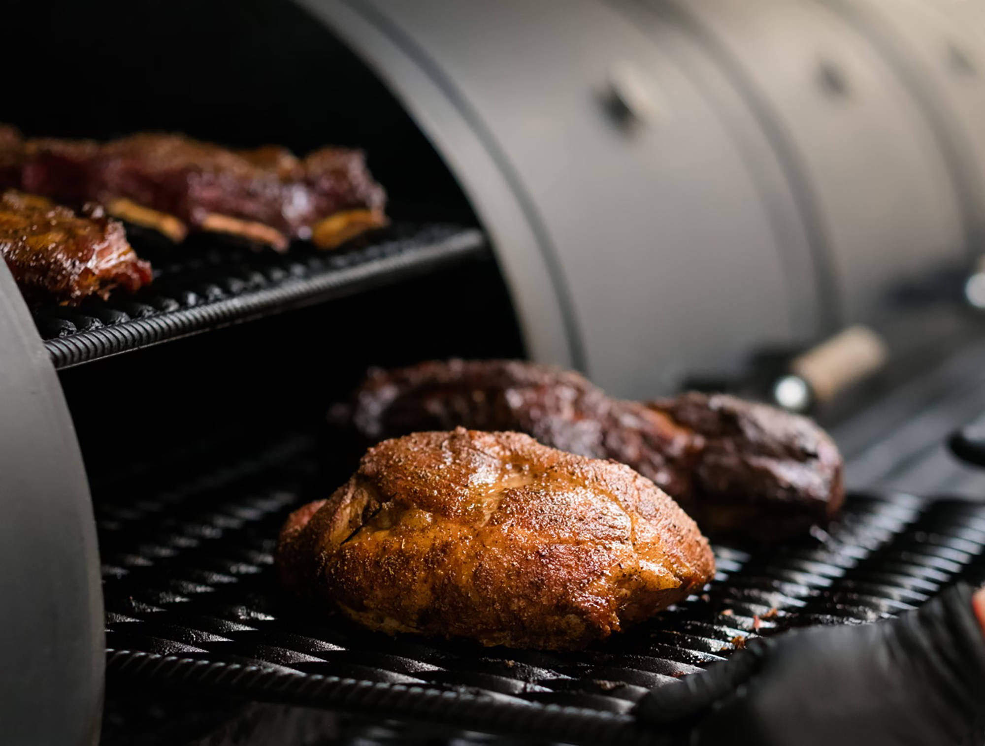 Großes Stück Fleisch mit Gewürzkruste im Smoker gegrillt, saftig gebräunt, weitere Fleischstücke auf oberem Rost.