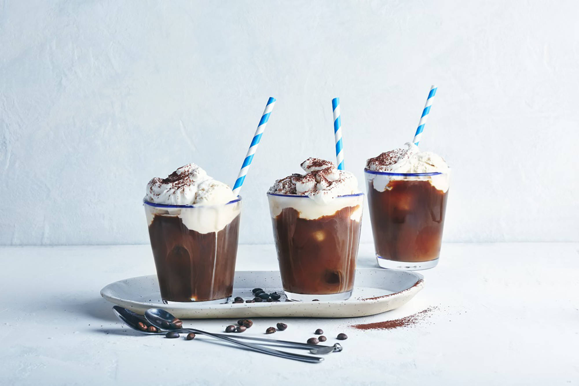 Drei Gläser Eiskaffee mit Schlagsahne, blauen Strohhalmen und Kakaopulver, serviert auf einem Tablett mit Kaffeebohnen.
