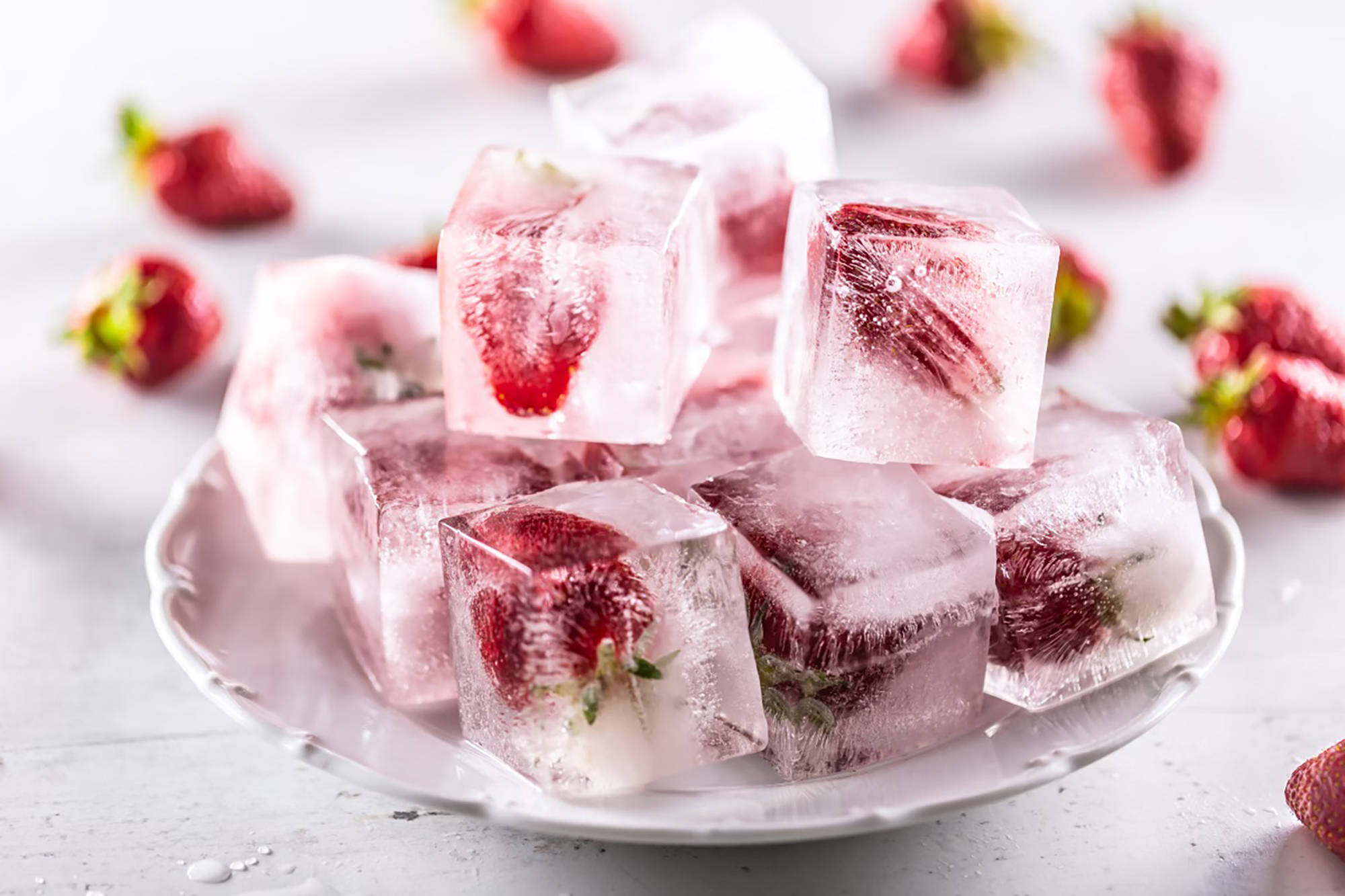 Eiswürfel mit eingefrorenen Erdbeeren auf einem weißen Teller, daneben frische Erdbeeren.