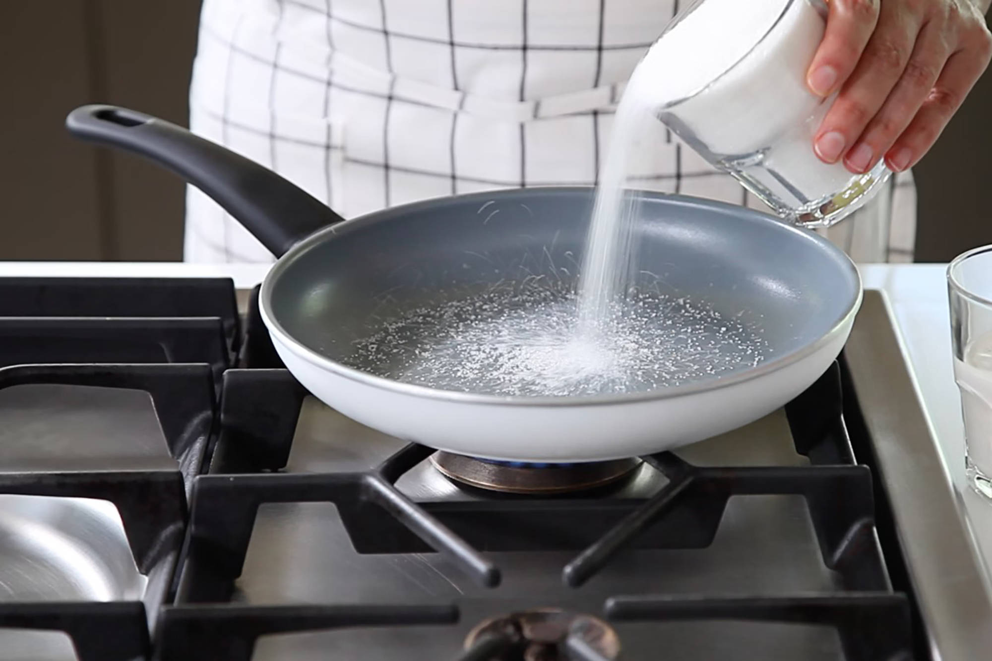 Person schüttet Zucker in eine Pfanne über einem Gasherd zum Karamellisieren.