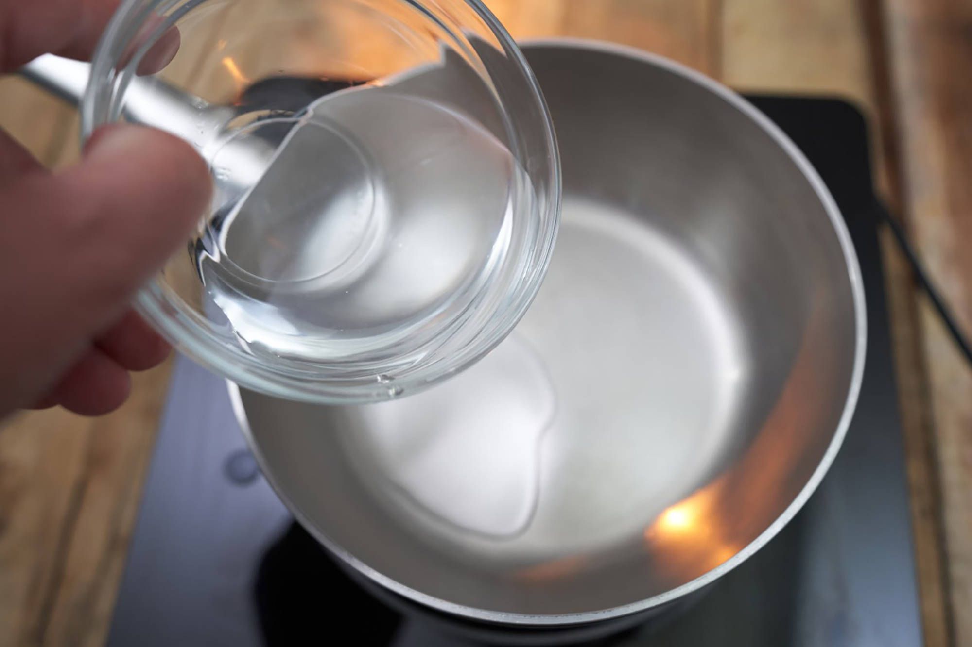Jemand gießt klares Wasser aus einer Glasschüssel in einen leeren Pfanne auf dem Herd.