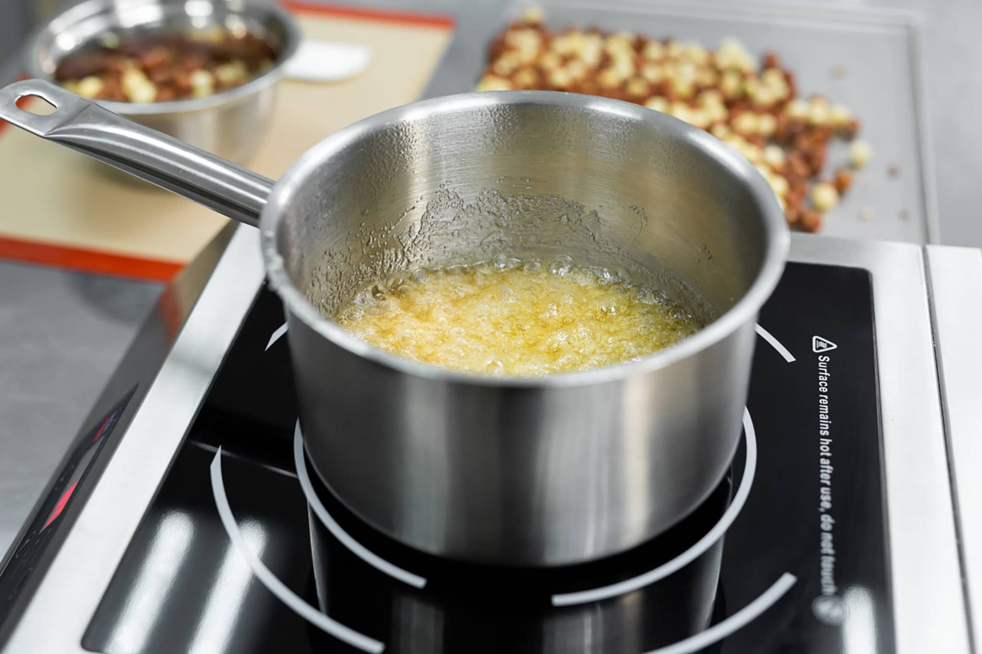 Karamellisieren von Zucker in einem Topf auf einem Herd, im Hintergrund geröstete Nüsse auf einem Blech.