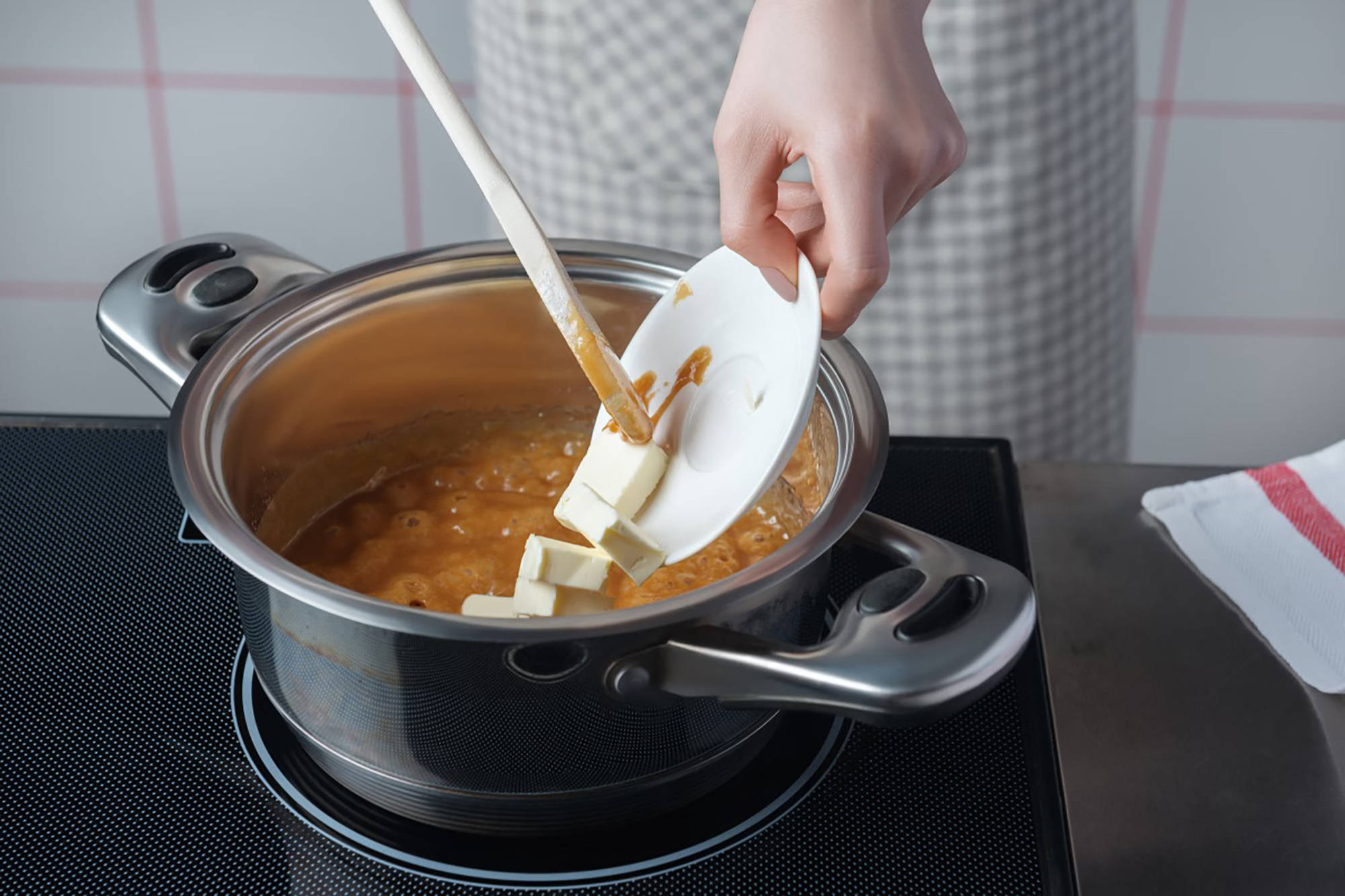 Eine Person gibt Butterstücke in einen Topf mit kochendem Karamell, während sie mit einem Holzlöffel rührt.