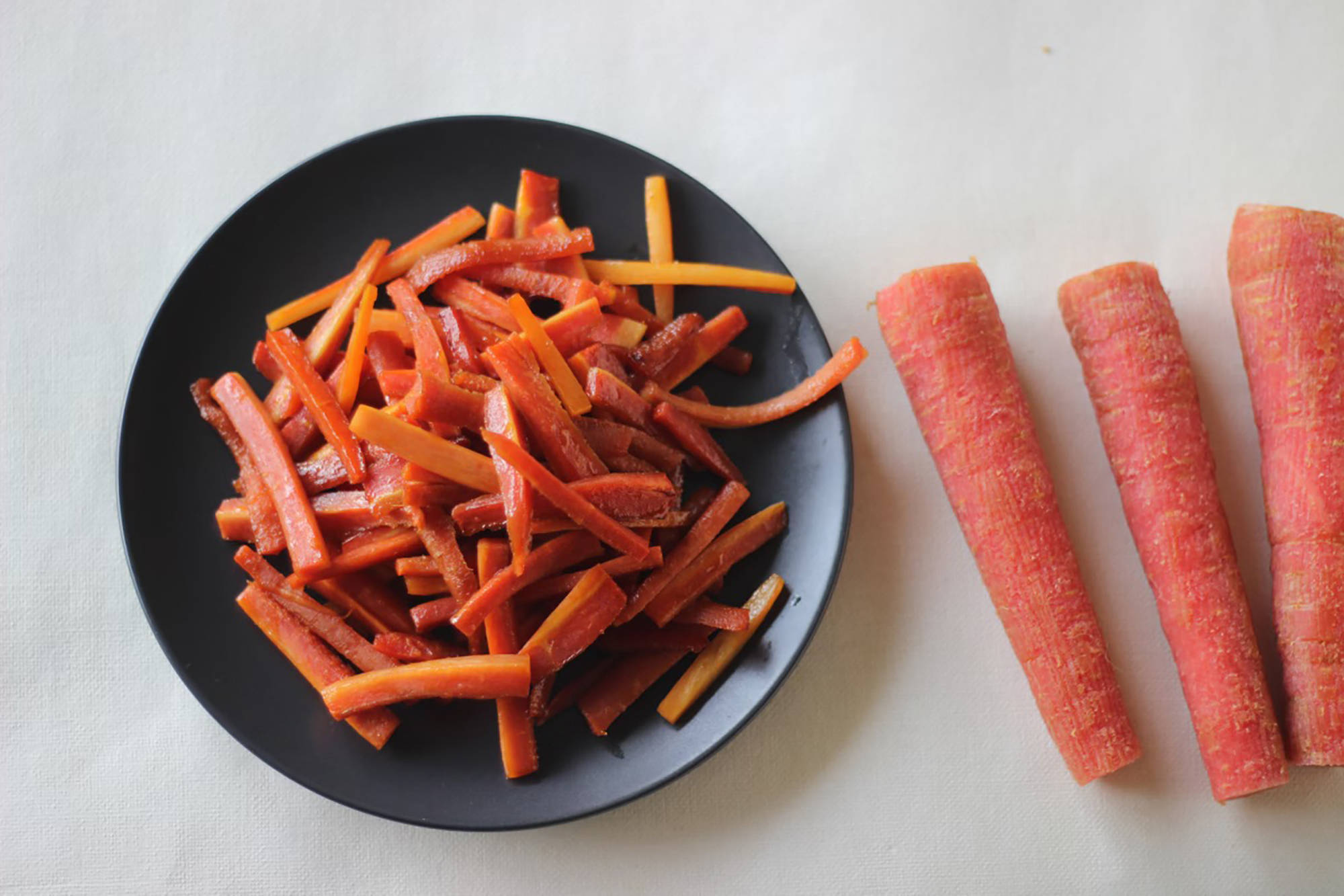 Geschnittene rote und orange Karottenstreifen auf einem schwarzen Teller, daneben liegen ungeschnittene Karotten auf ein