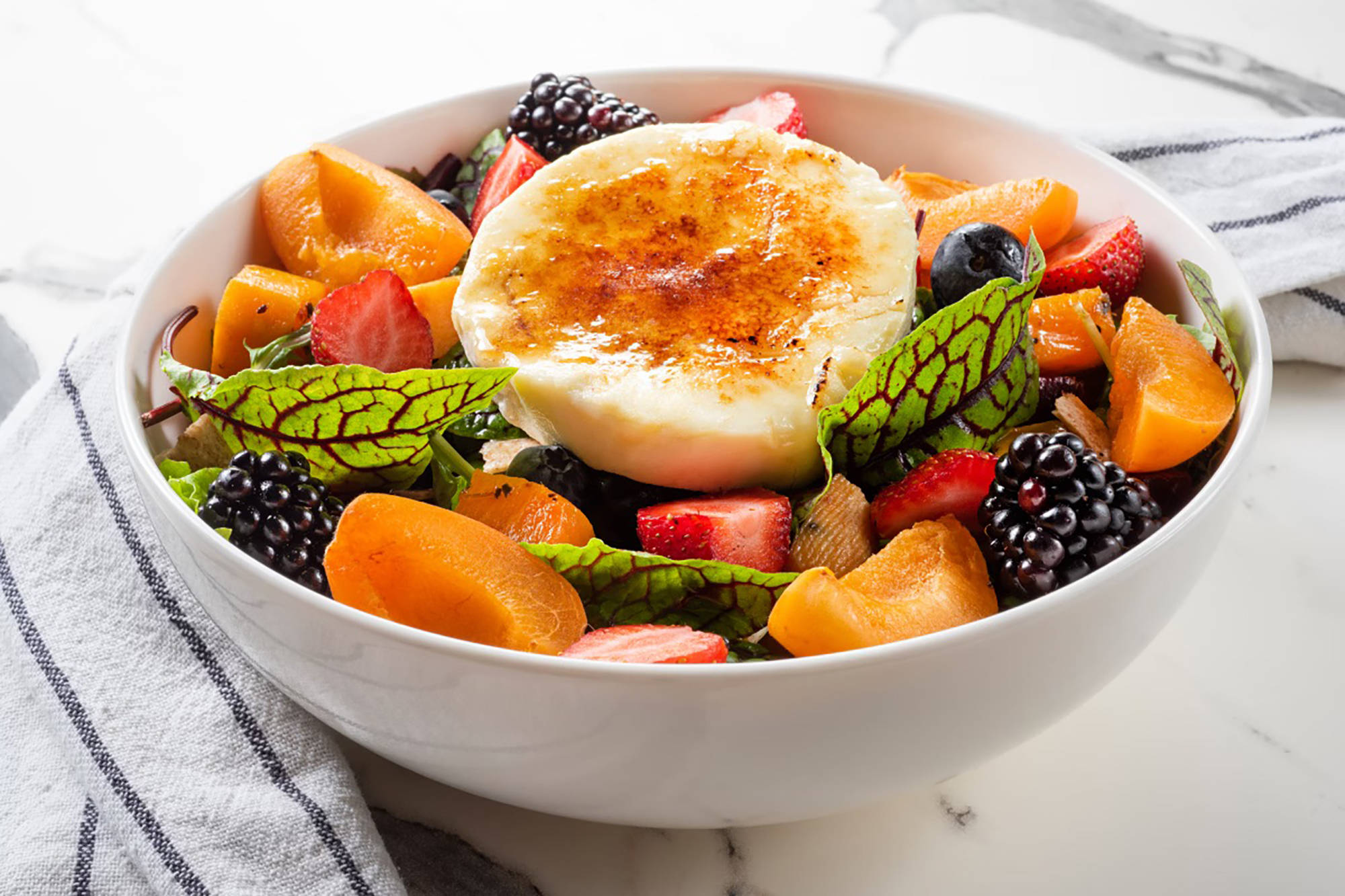 Frischer Salat mit karamellisiertem Ziegenkäse, Brombeeren, Erdbeeren und Aprikosen in einer weißen Schüssel serviert.