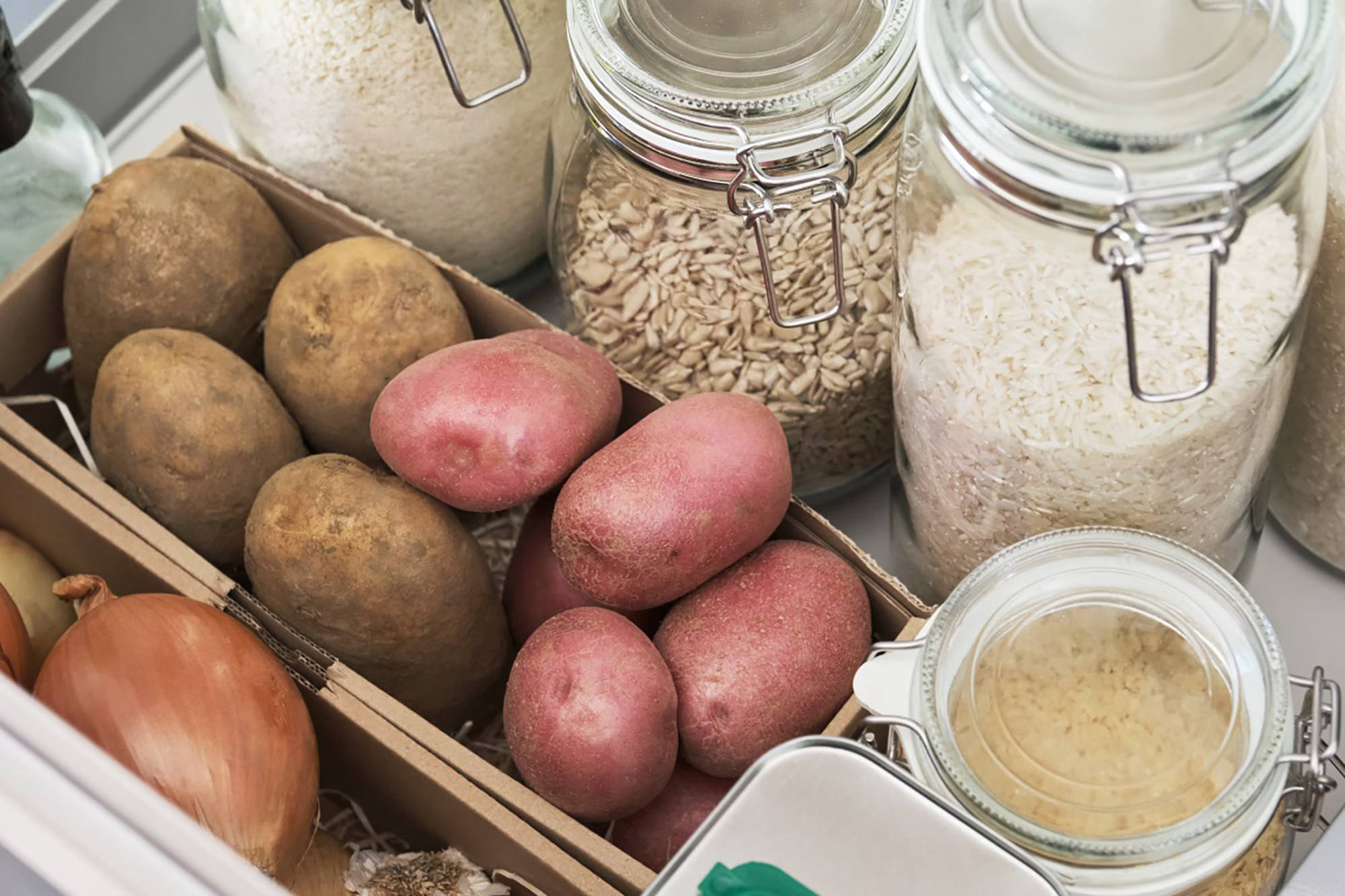 Kartoffeln lagern in einem Karton, umgeben von Vorratsgläsern mit Reis und Getreide.