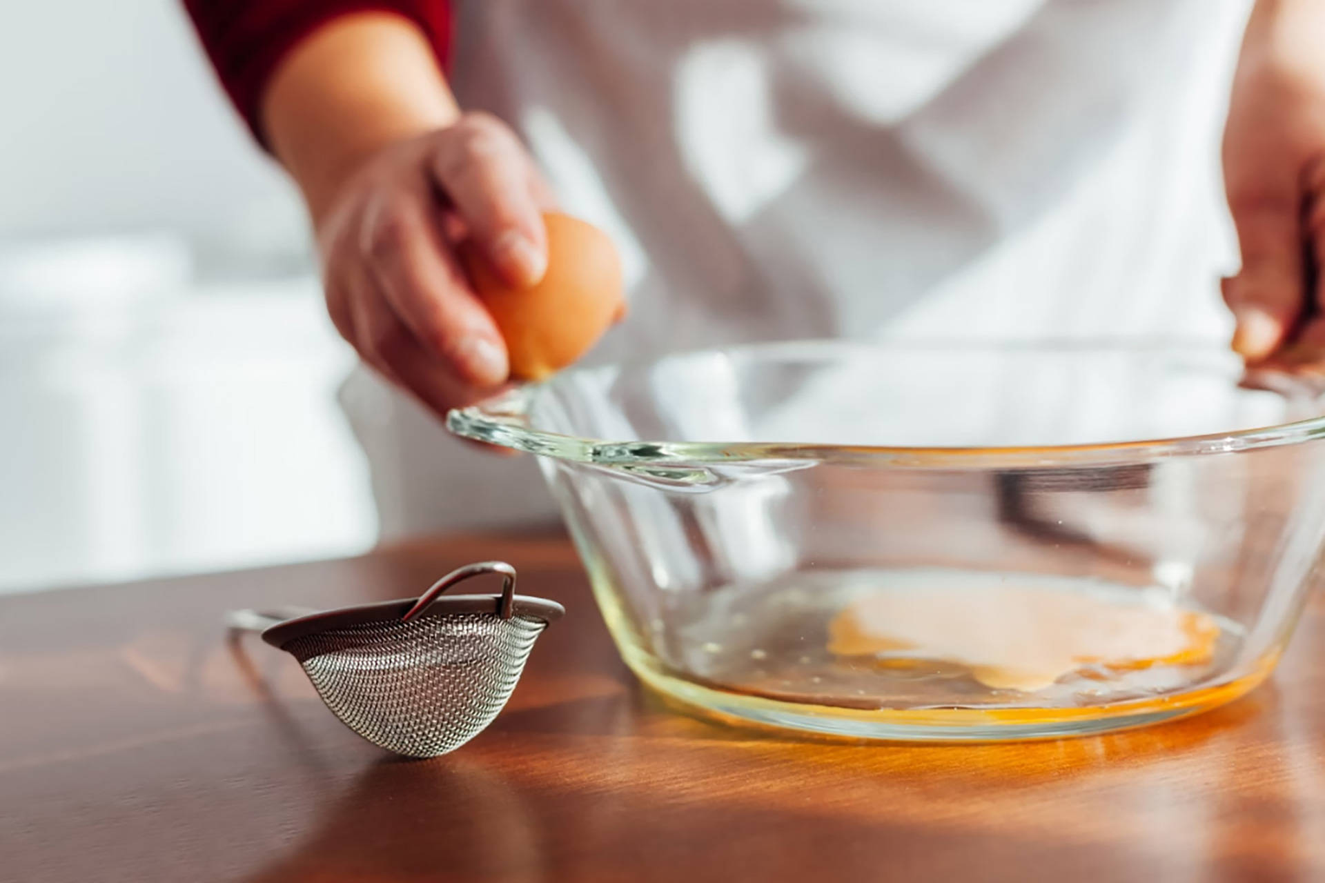 Person trennt Ei in Glasschüssel, Sieb auf Tisch.