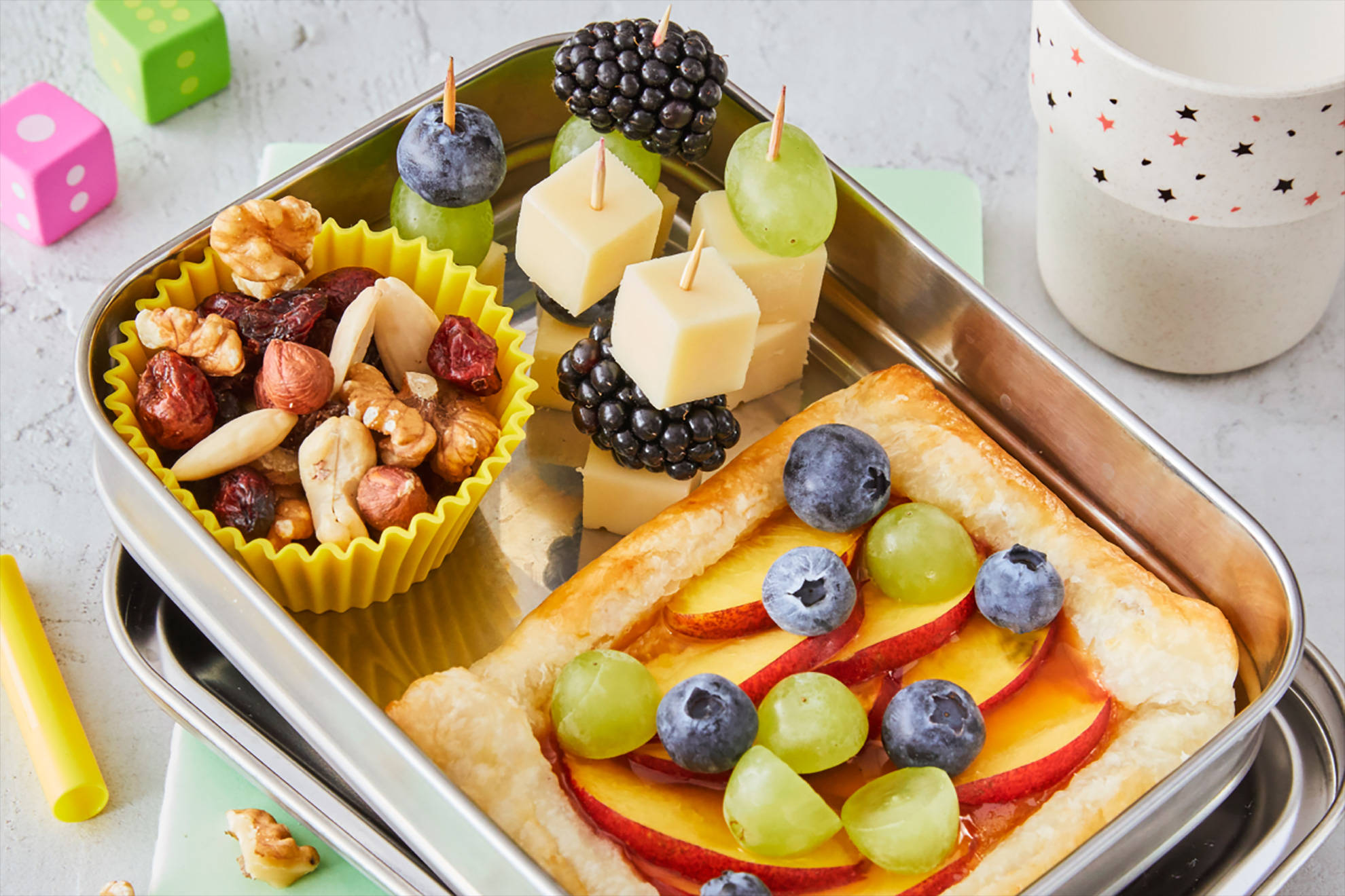 Eine Aluminium-Brotdose, gefüllt mit kleinen Käse-Beeren-Spießen, Nüssen und einer gesunden Obst-Pizza.