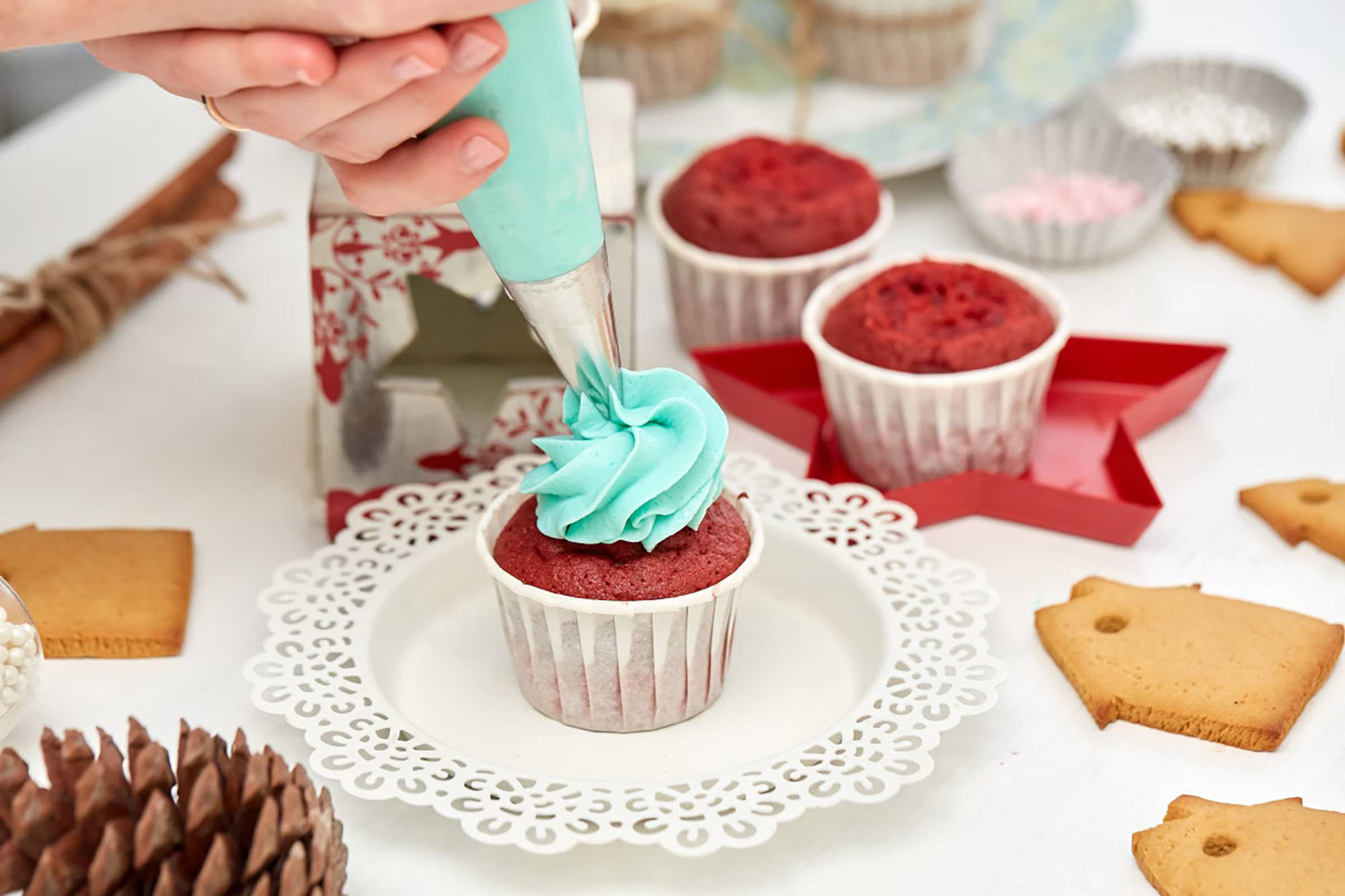 Jemand verziert mit einem Spritzbeutel einen roten Cupcake mit hellblauem Frosting auf dekorativem Teller.