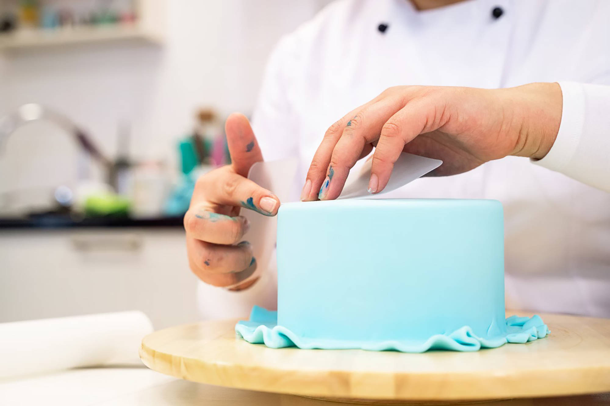 Jemand glättet die Oberfläche einer blauen Fondanttorte mit einem Glättwerkzeug, die auf einem Holzdrehteller steht.