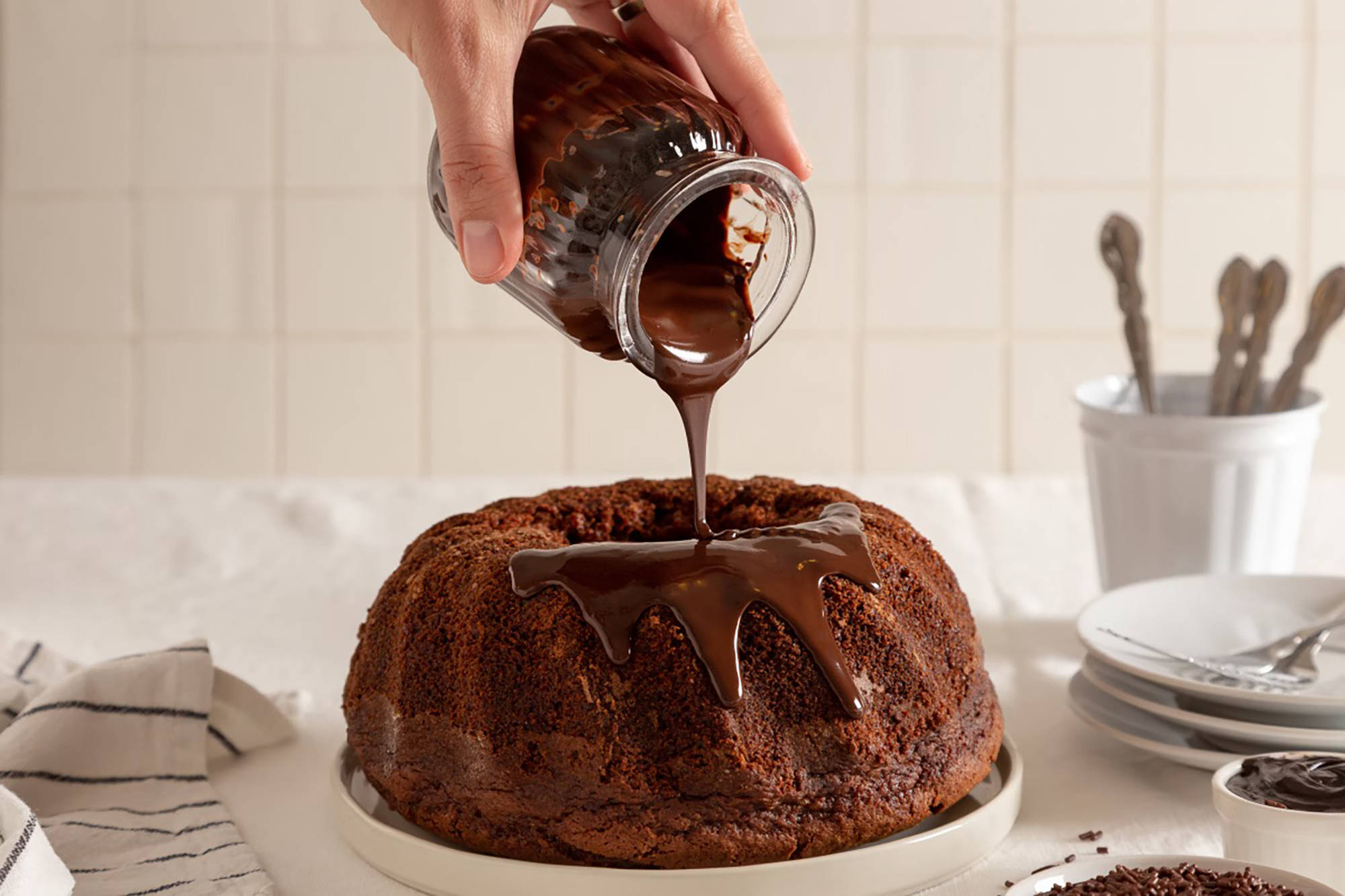 Jemand gießt flüssige Schokolade auf einen Schokoladen-Gugelhupf der auf einem Teller steht.