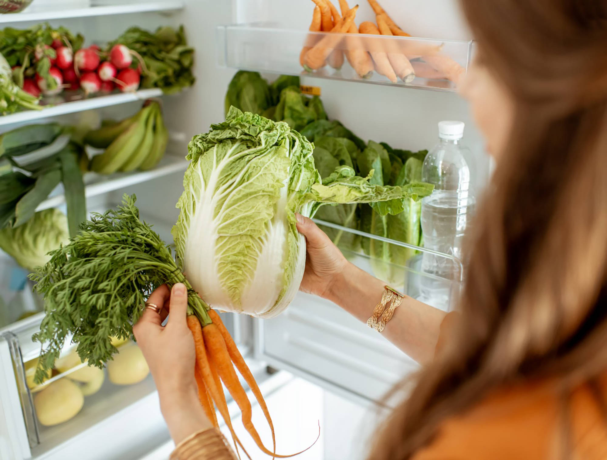 Frau räumt frisches Gemüse, darunter Chinakohl und Karotten, in einen gut gefüllten Kühlschrank mit gesunden Lebensmitte