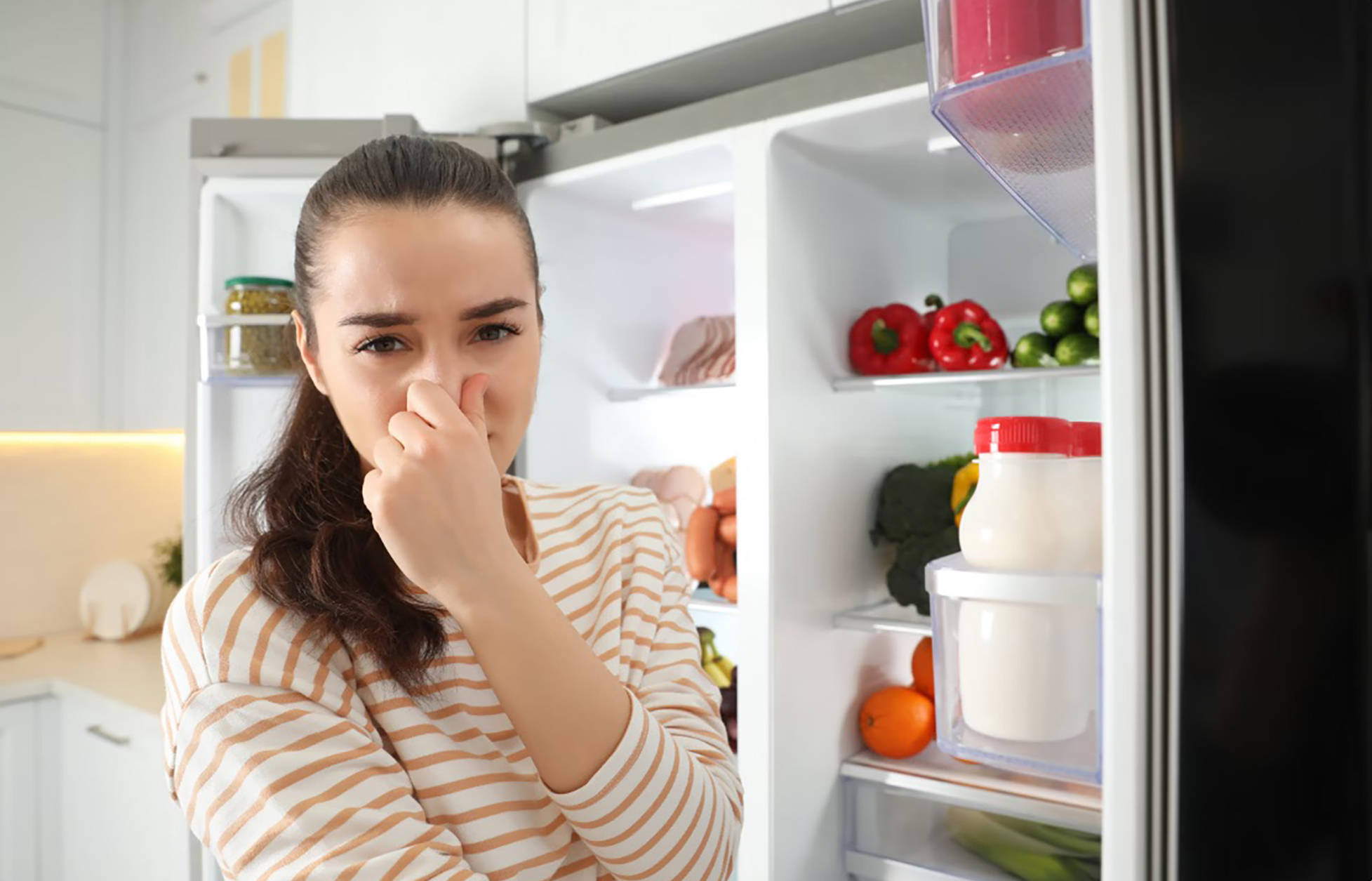 Frau hält sich die Nase zu, während sie vor einem offenen Kühlschrank mit frischen Lebensmitteln steht.