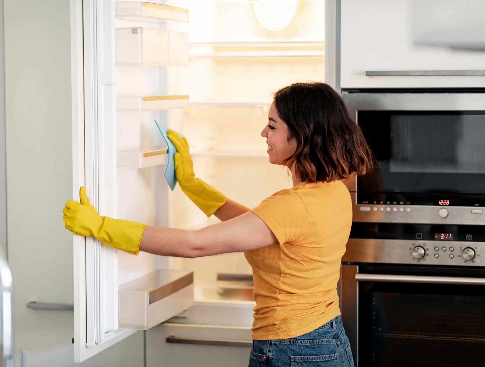 Frau mit gelben Gummihandschuhen reinigt die Tür eines leeren Kühlschranks mit einem blauen Tuch in einer modernen Küche
