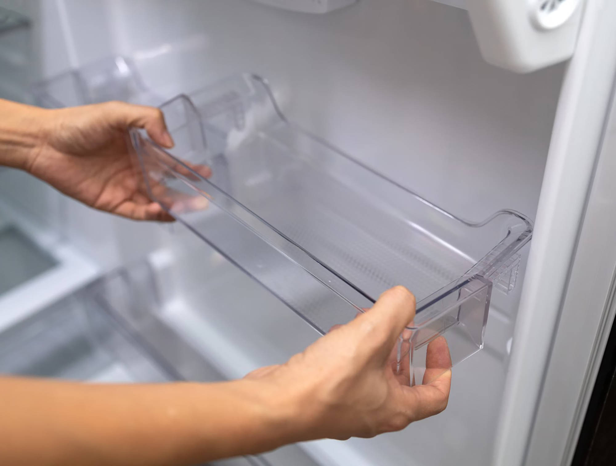 Nahaufnahme von Händen, die ein transparentes Kunststofffach in die Tür eines leeren Kühlschranks einsetzen oder es zur 