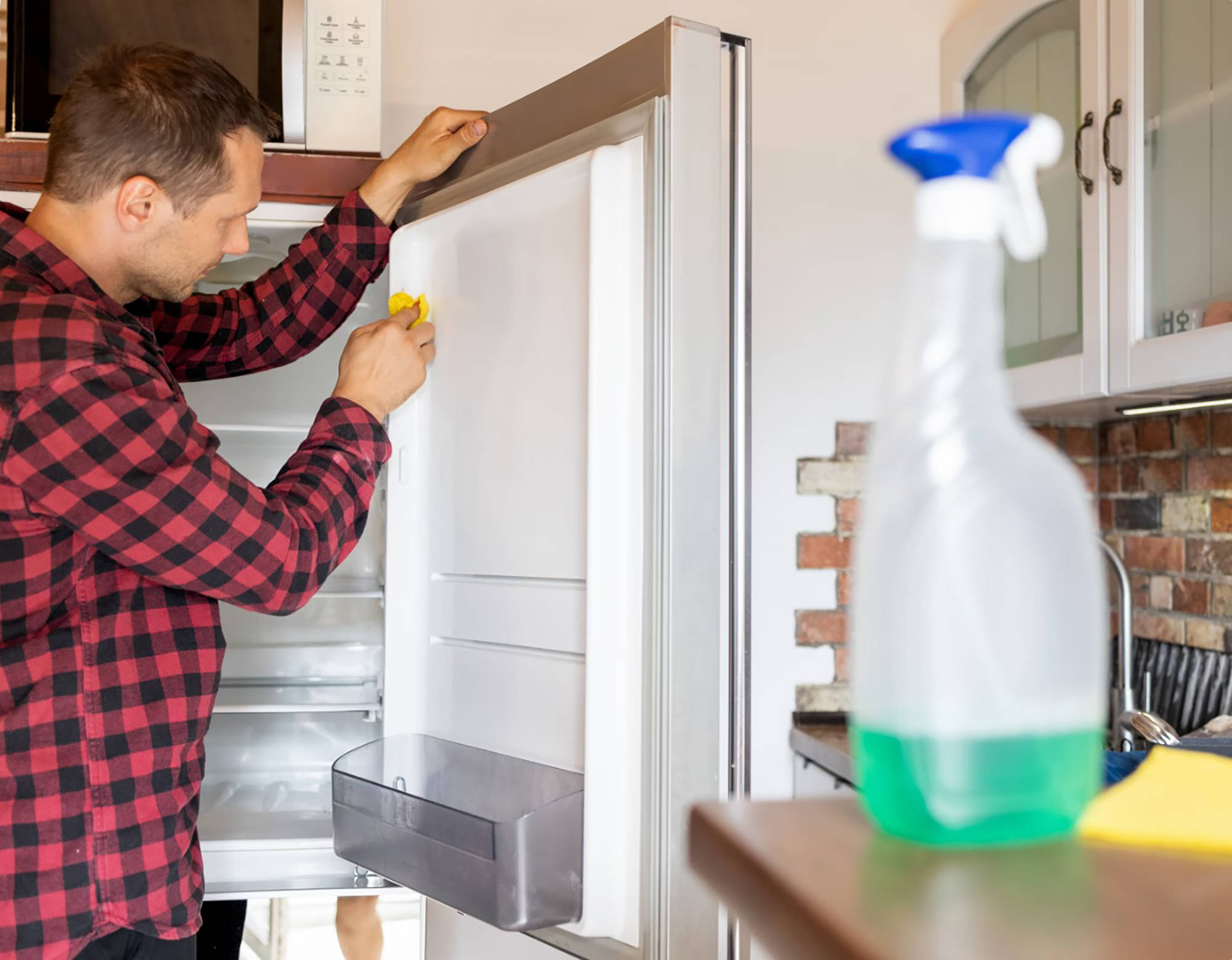 Mann in kariertem Hemd reinigt die Türdichtung eines offenen Kühlschranks mit einem gelben Tuch in einer rustikalen Küch
