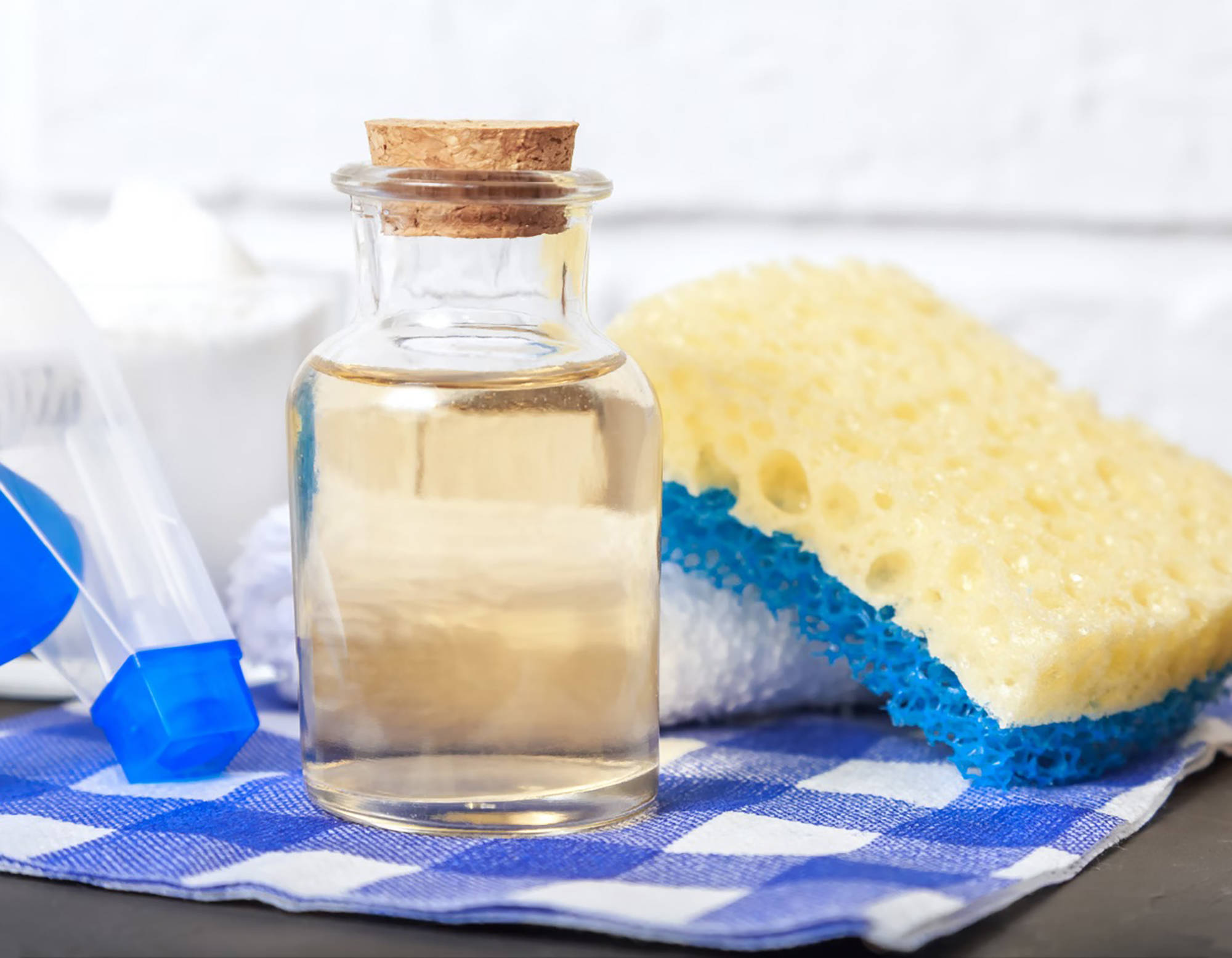Glasflasche mit Essig, gelber Schwamm, Sprühflasche und Natron auf blau-weiß kariertem Tuch.