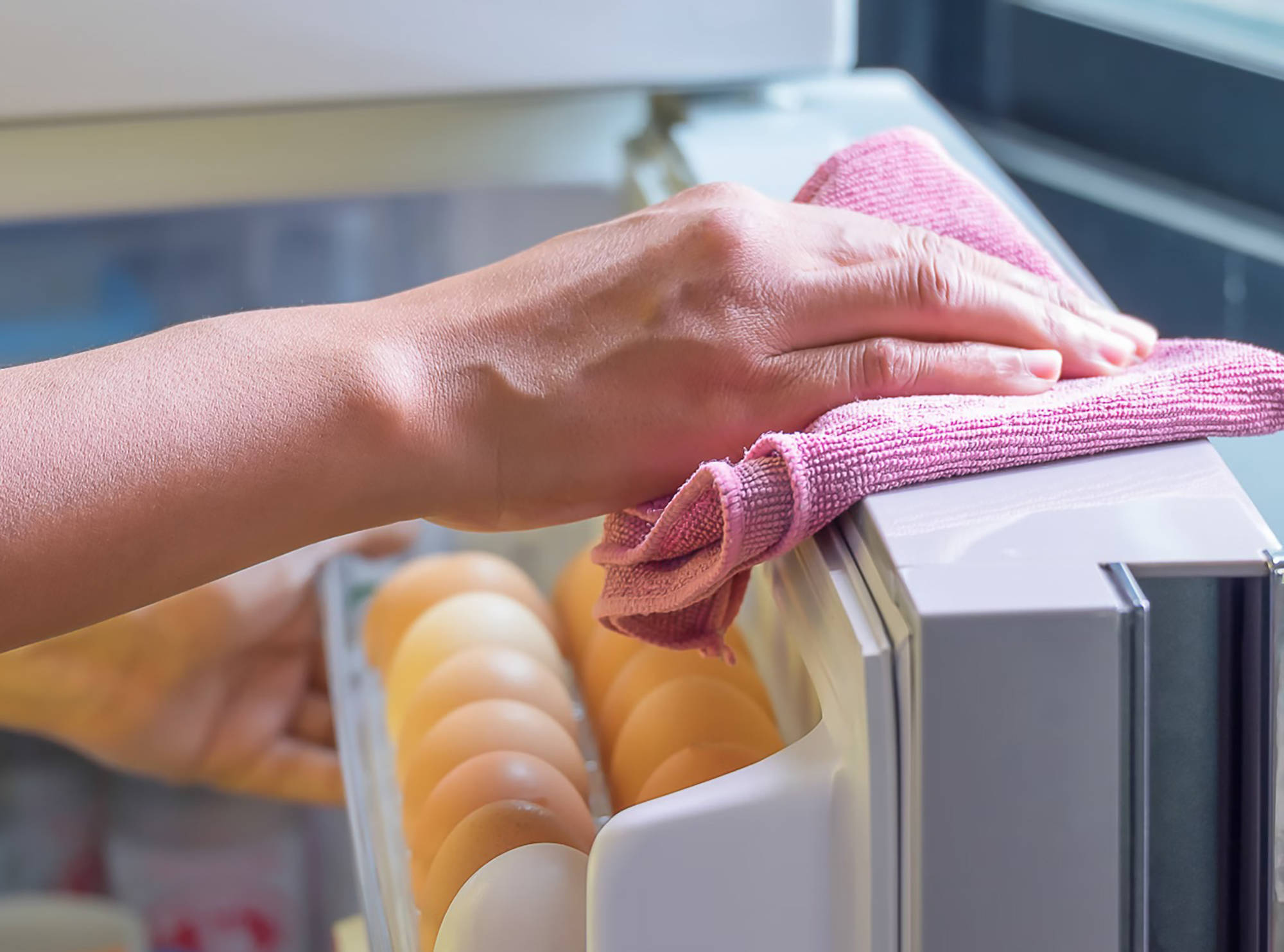 Eine Person wischt mit rosa Mikrofasertuch die Oberseite eines Kühlschranks neben einem Eierfach sauber.