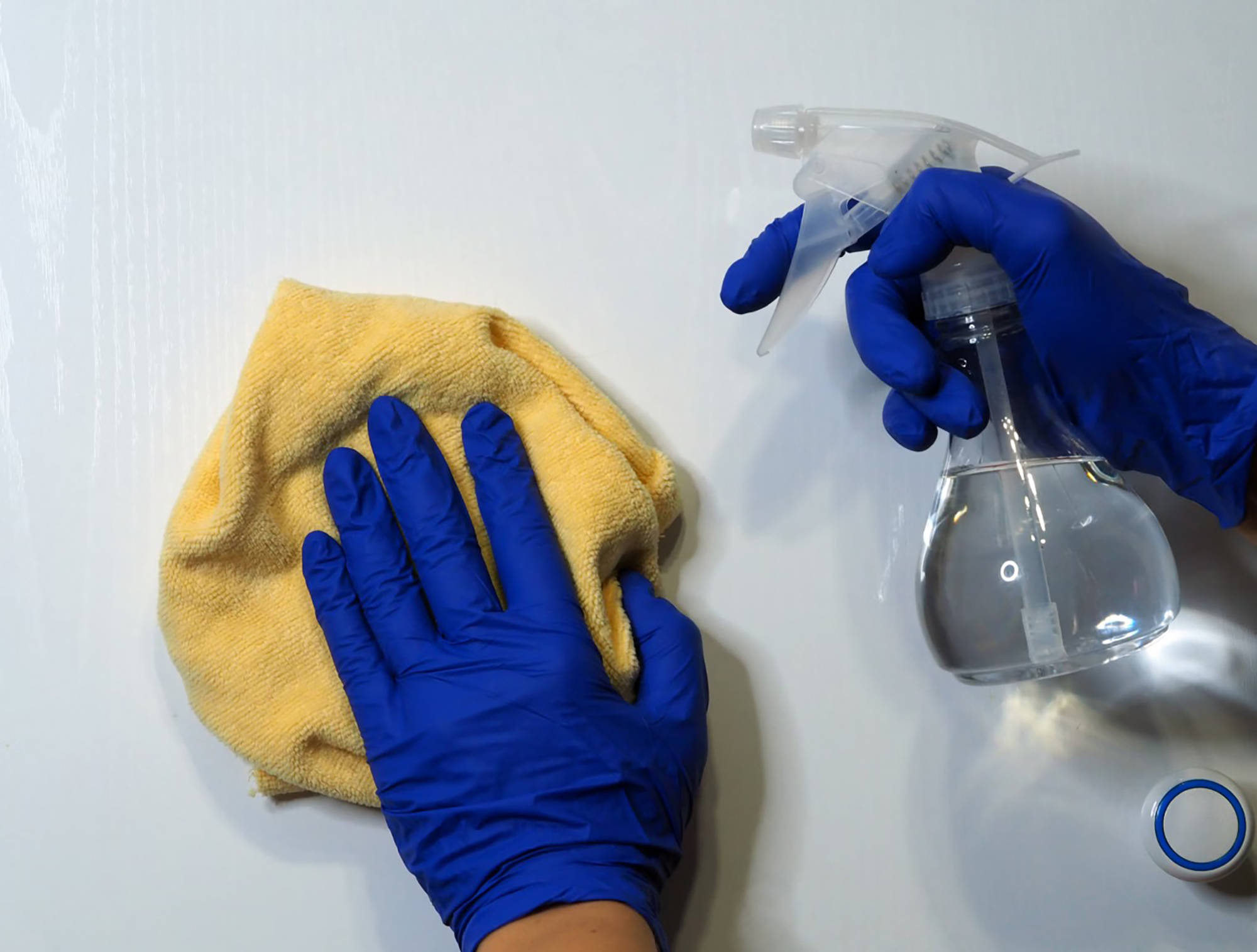 Person mit blauen Handschuhen reinigt eine Oberfläche mit gelbem Mikrofasertuch und transparenter Sprühflasche.