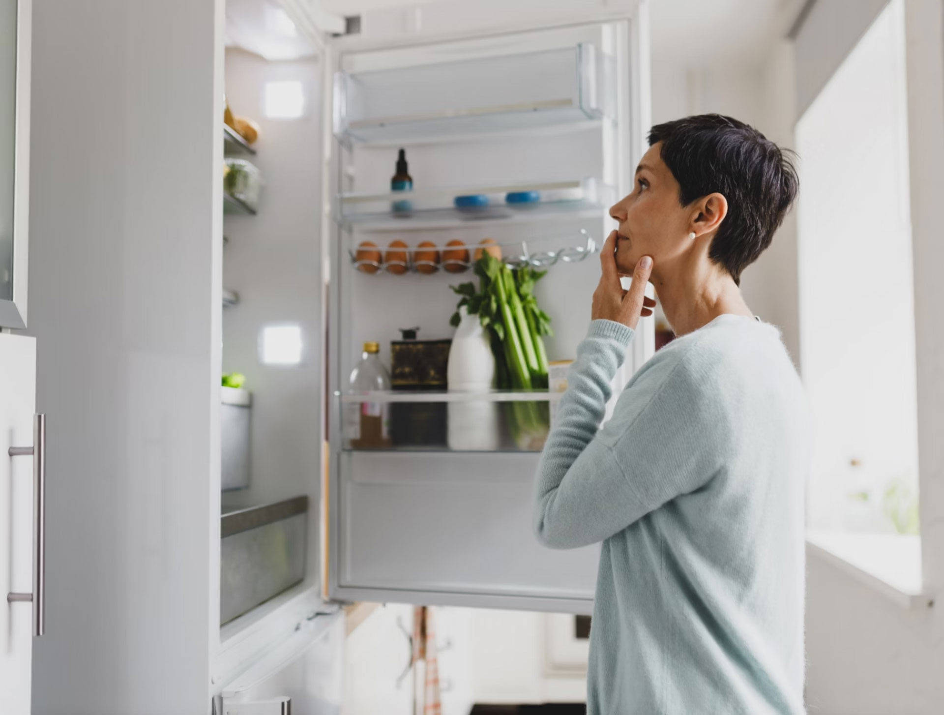 Frau in hellblauem Pullover schaut in einen offenem Kühlschrank gefüllt mit Eiern, Gemüse und Flaschen in einer hellen K