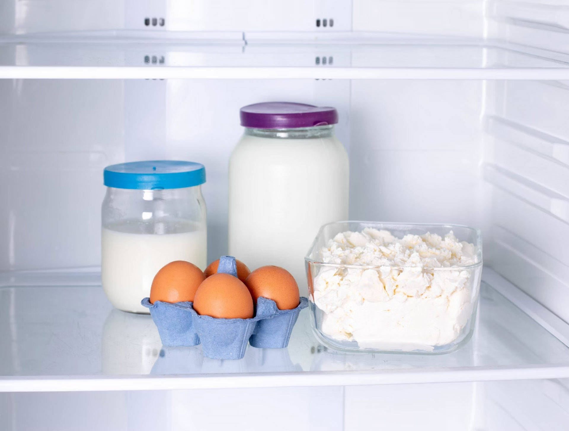 Kühlschrankfach mit Eiern in blauer Verpackung, zwei Milchgläsern und einer Glasschüssel mit frischem Hüttenkäse.