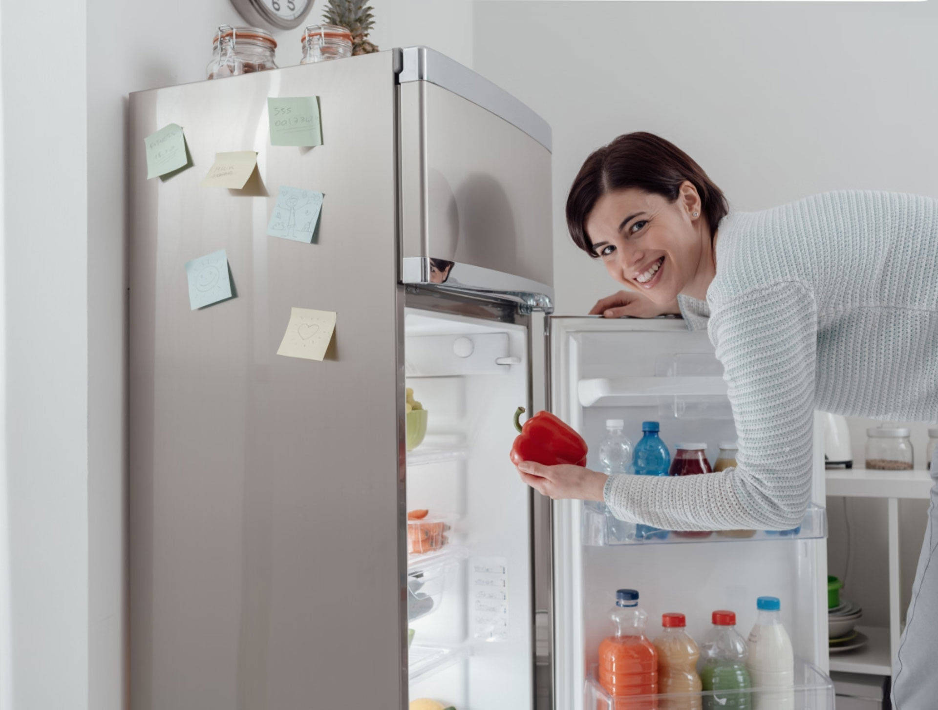 Lächelnde Frau holt eine rote Paprika aus einem offenen silbernen Kühlschrank.