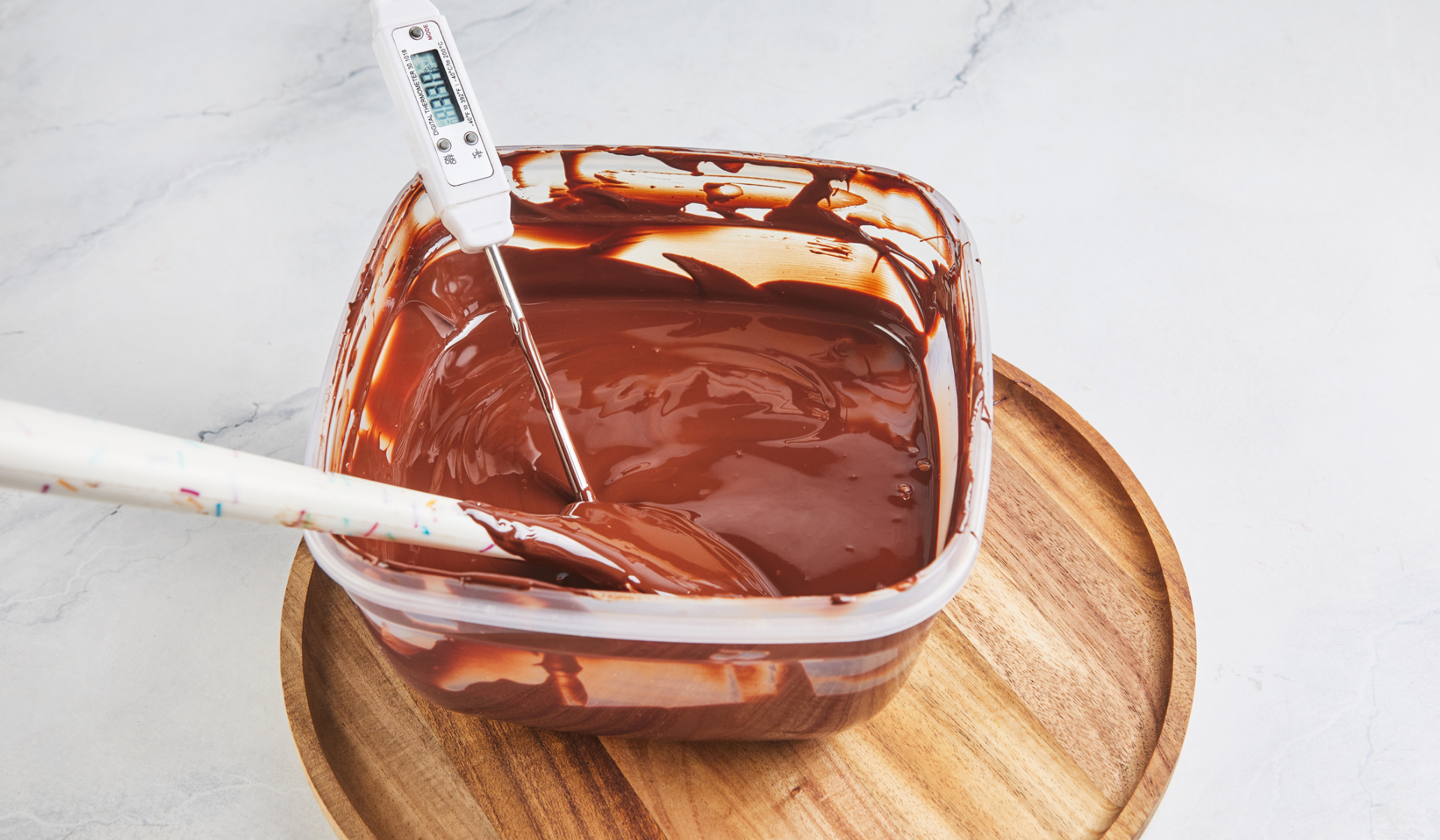 Eine Plastikbox mit geschmolzener Kouvertüre und einem Holzlöffel und einem Thermometer.