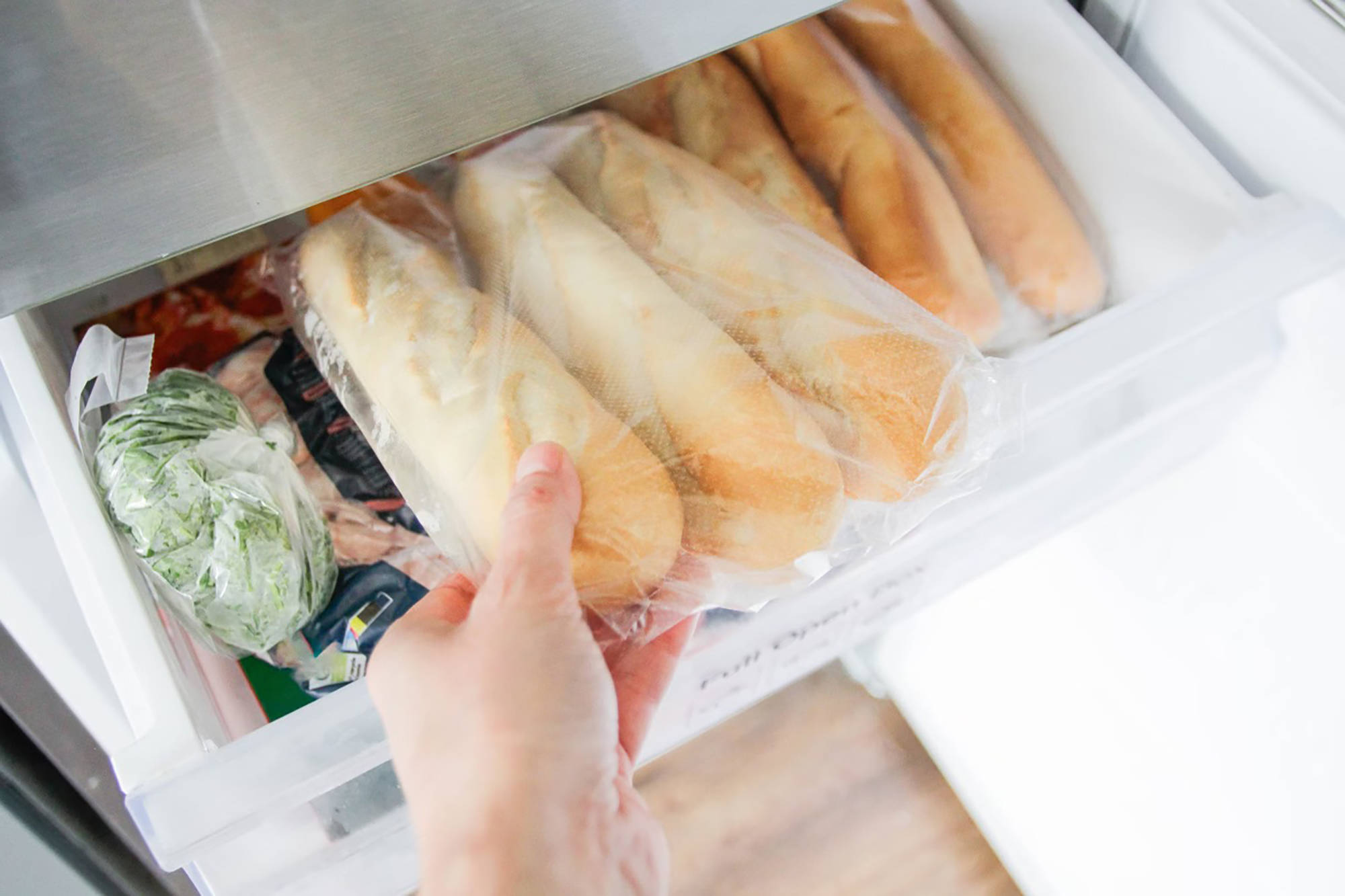 Jemand legt in Folie verpackte Baguettes ins Gefrierfach neben anderen tiefgefrorenen Lebensmitteln.