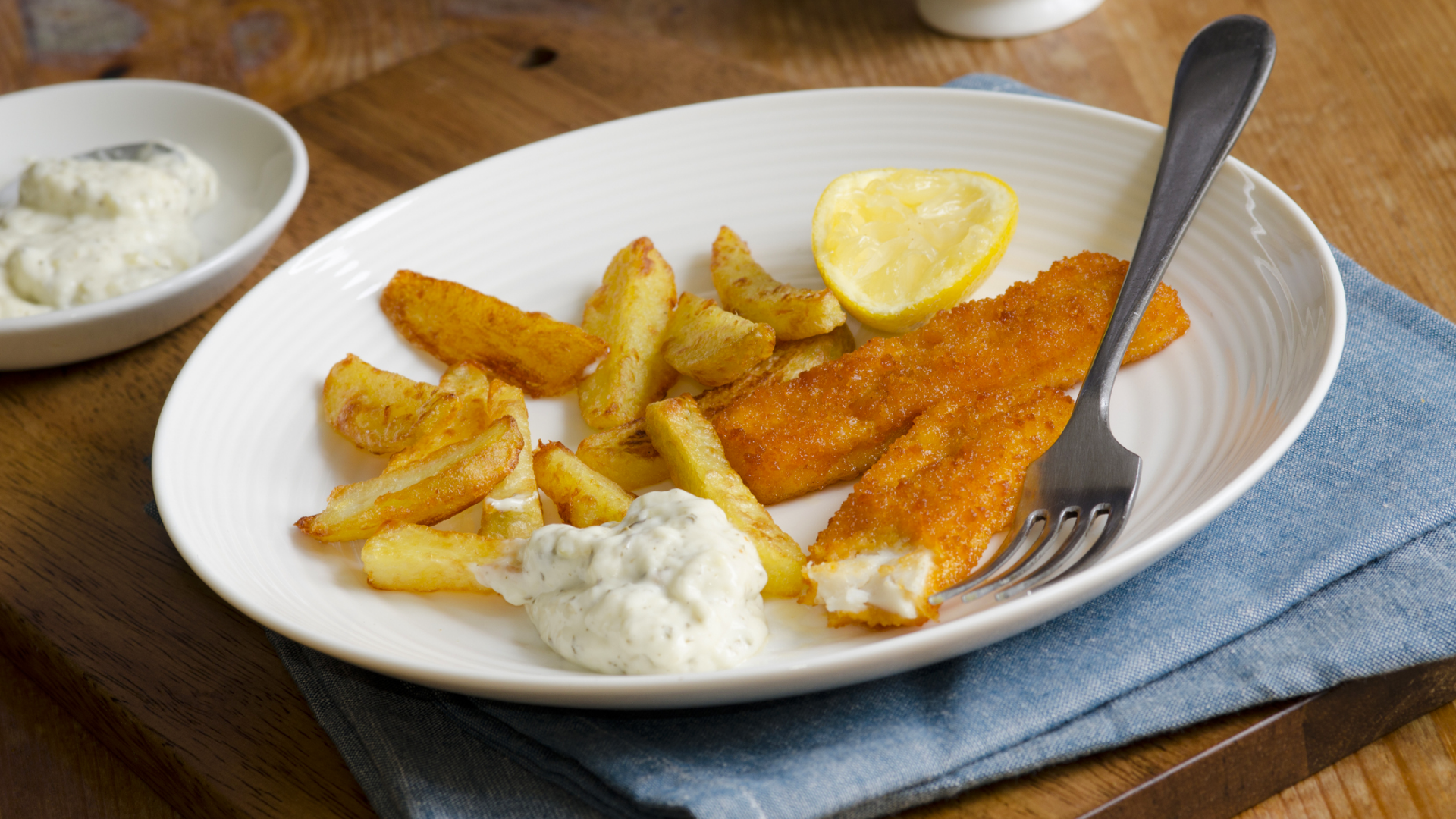 Panierter Fisch, Kartoffelspalten, Zitrone, Remoulade auf Teller.